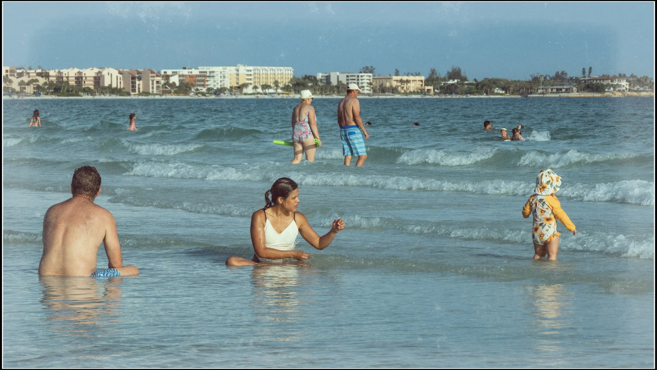 People enjoying a day at the beach with ocean waves, some in the water and others sitting or wading near the shore, with buildings in the background.