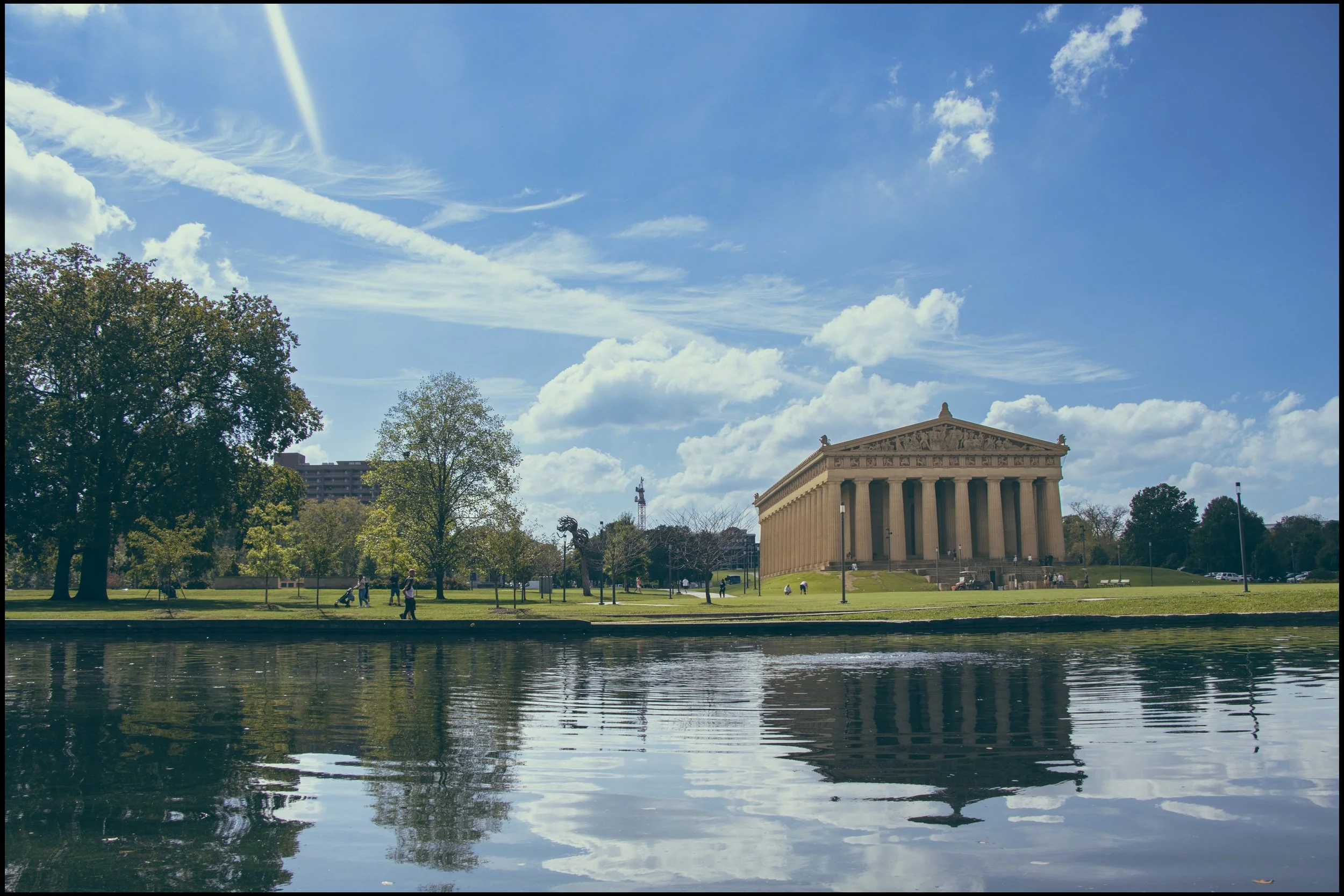 The Lincoln Memorial is seen from across a reflecting pool in a park, with trees and people walking along the grass under a partly cloudy sky.