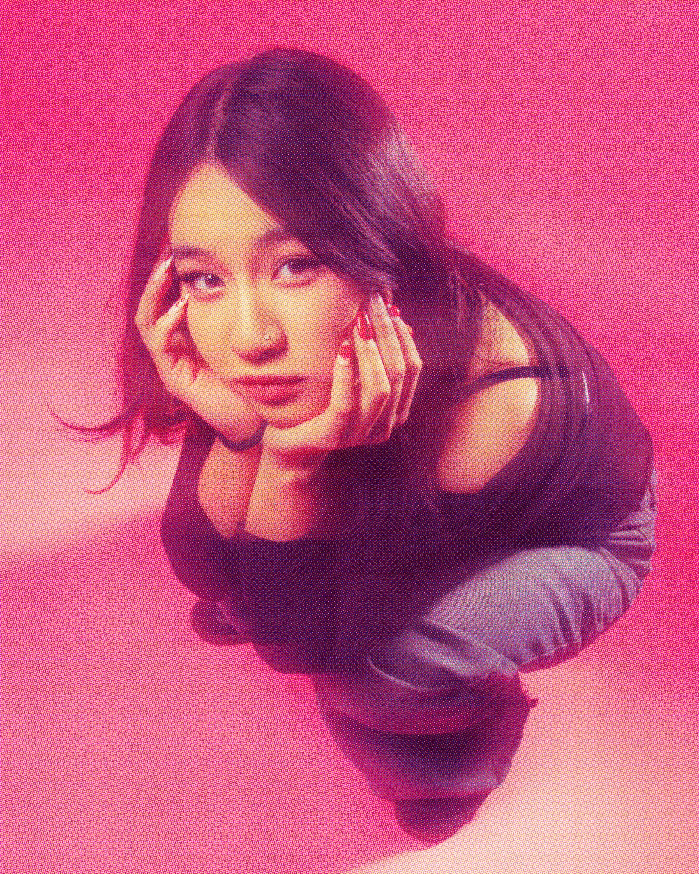 A woman with dark hair styled in loose waves, wearing a black top, is resting her chin on her hands, with a pink gradient background.