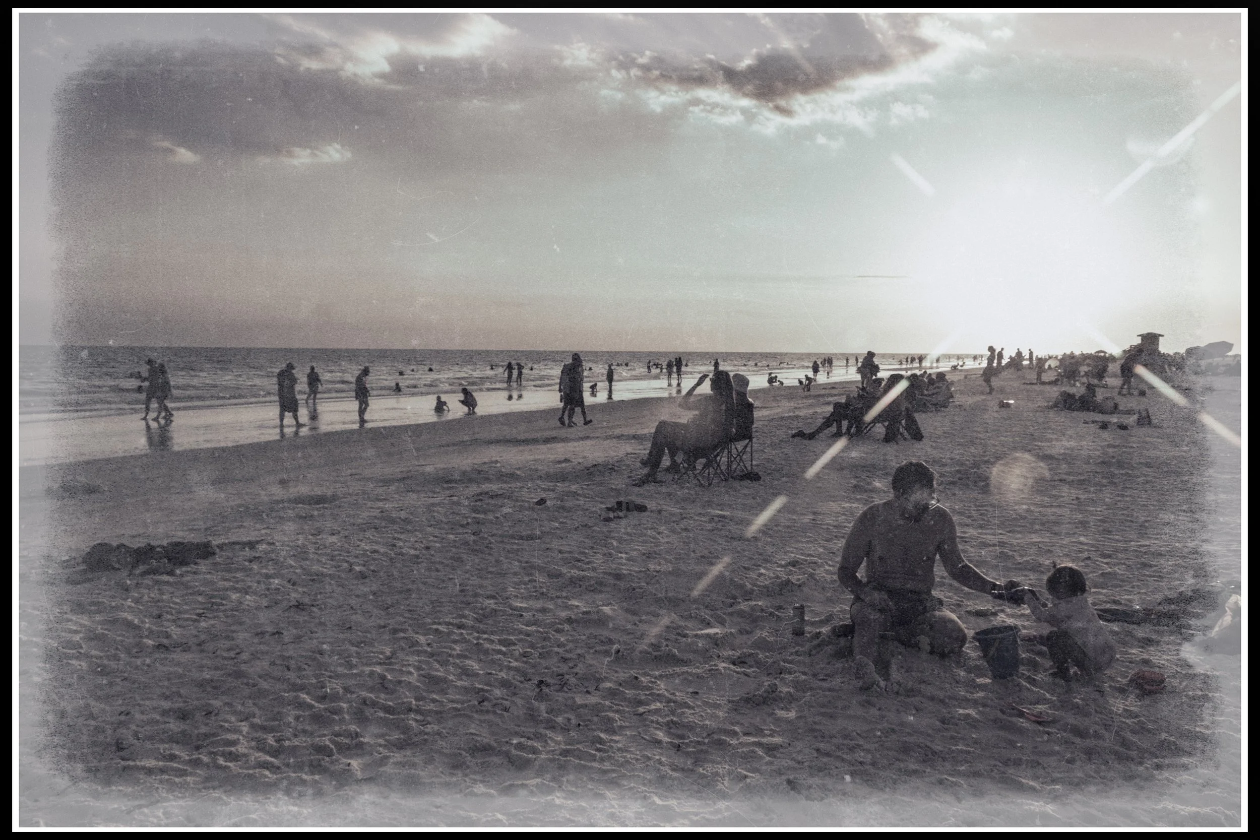 People on a beach during sunset with calm waves, some sitting in chairs, playing, and walking, under a cloudy sky.