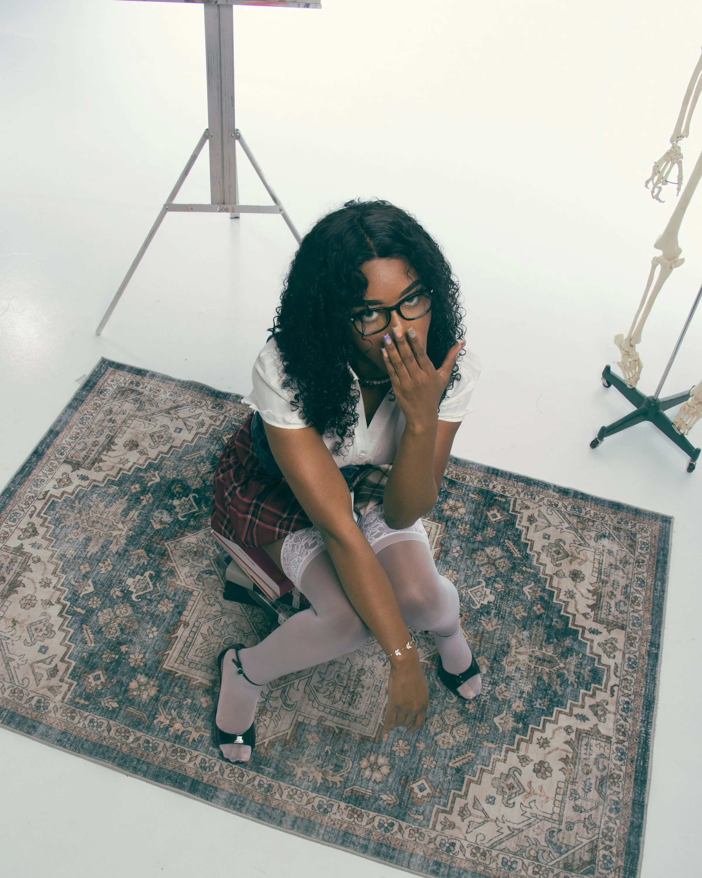 Young woman with curly black hair and glasses kneeling on a patterned rug in front of a white background. She is wearing a white top, plaid skirt, white tights, and black shoes, and is covering her mouth with her hand. There is a treadmill and a skel