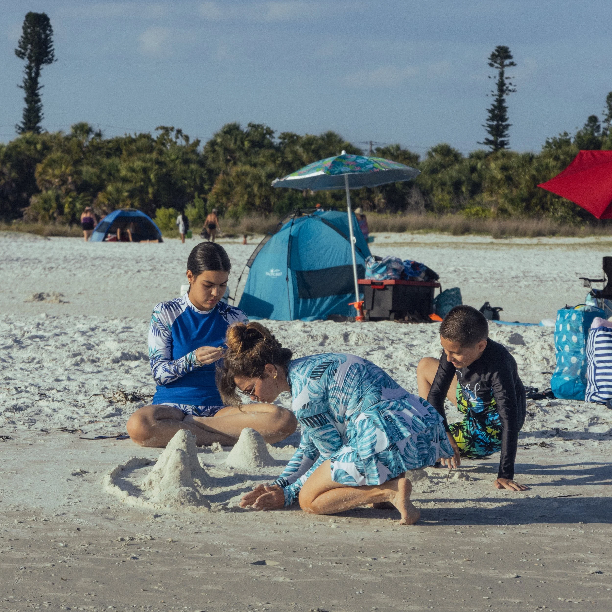 People practicing yoga outdoors on a sandy beach with tents, umbrellas, trees, and a cloudy sky in the background.