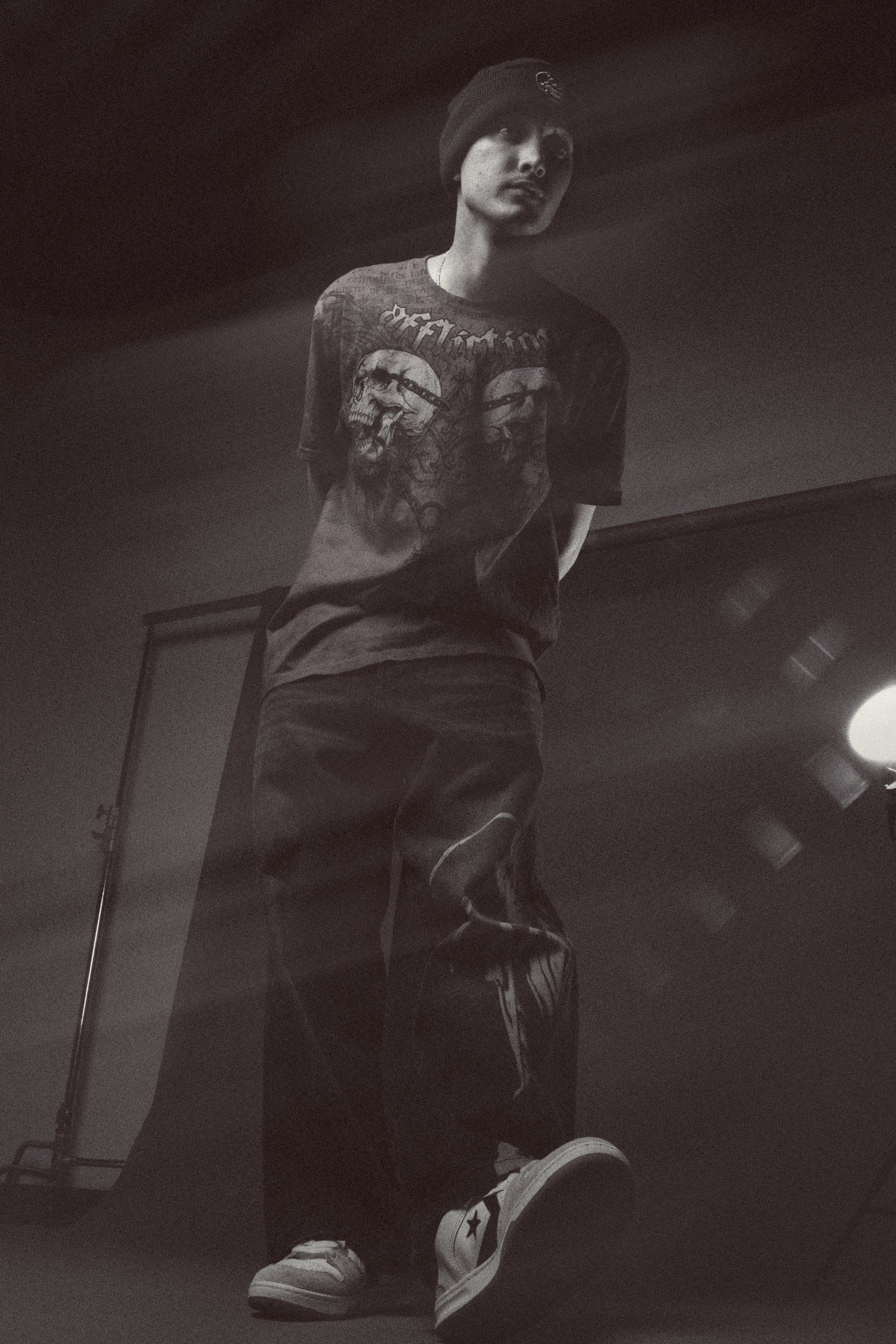 A person standing indoors, wearing a black beanie, dark graphic T-shirt featuring a skull and text, dark pants, and white sneakers with a star logo, with a mirror and light fixture visible in the background.
