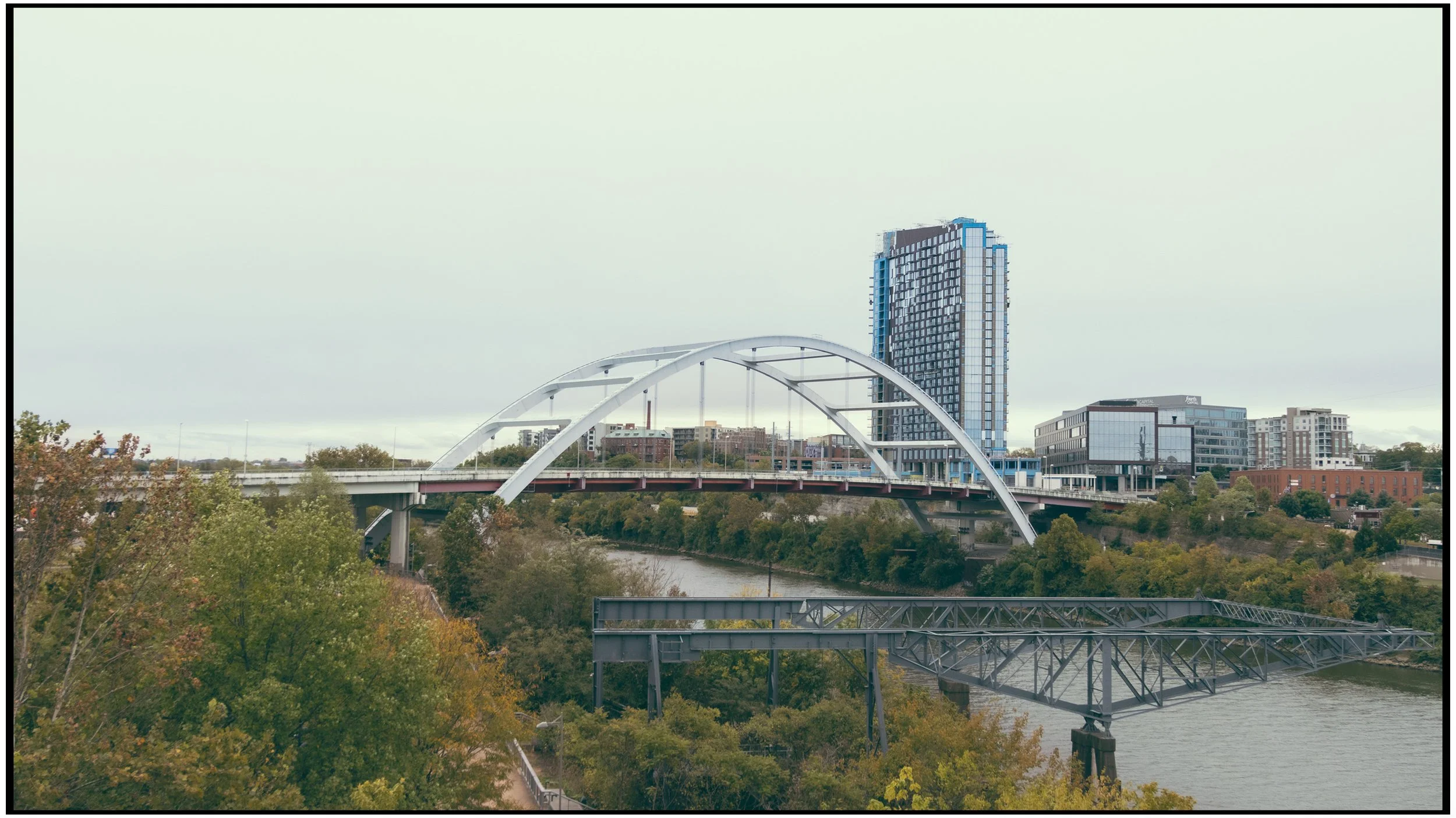 An urban cityscape showing a modern high-rise building, a bridge with white arches spanning a river, and surrounding greenery.