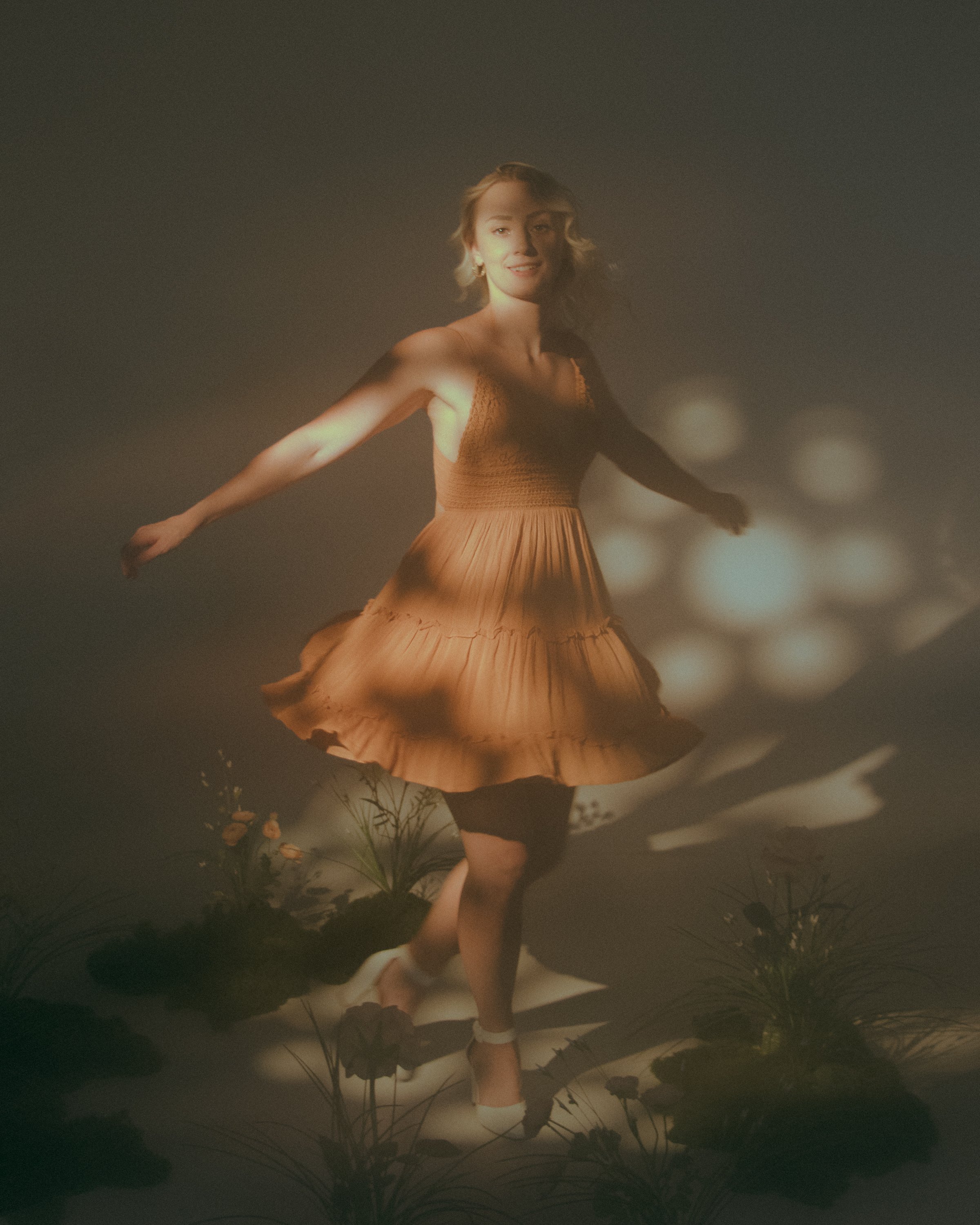 A woman dancing in a dress, with arms outstretched, on a stage with greenery and flowers in the foreground, illuminated by soft, warm lighting.