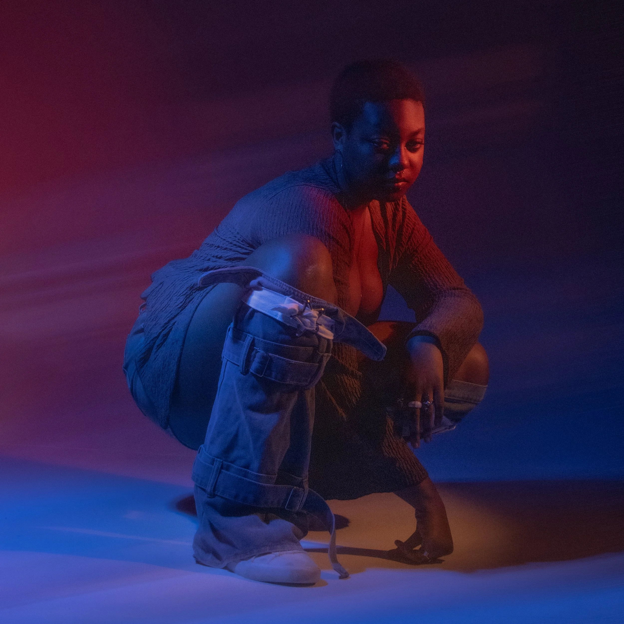 A person with short hair and earrings, sitting with one knee up and arms wrapped around it, in front of a gradient background of blue, purple, and red lighting.