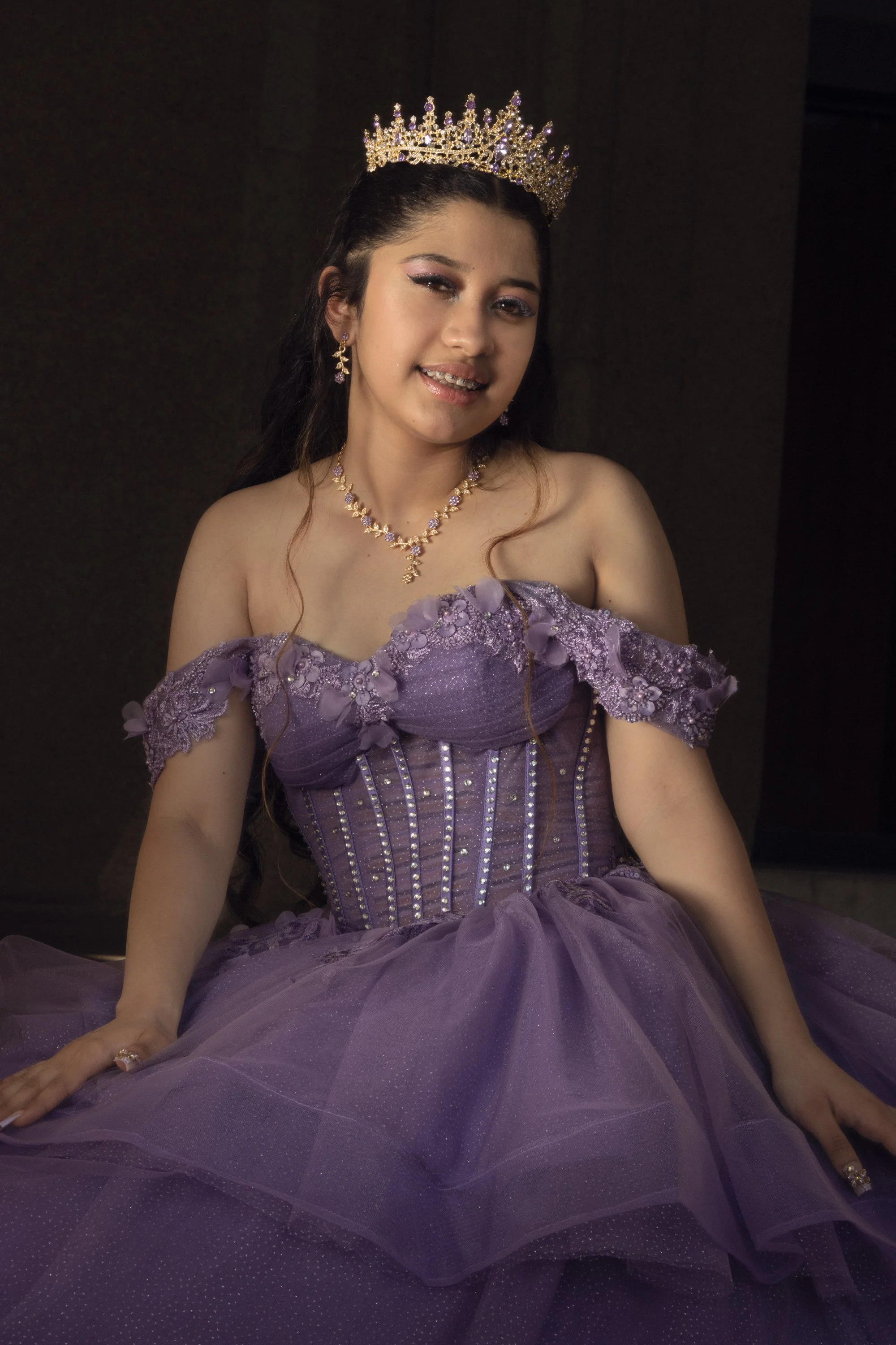 A young woman wearing a purple ball gown with floral embroidery and rhinestones, accessorized with a crown, earrings, a necklace, and a bracelet, sitting against a dark background.