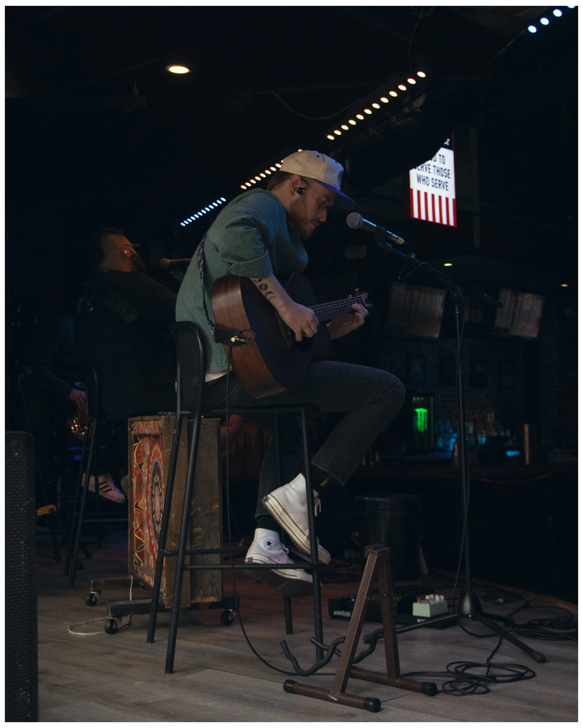 A man wearing a white cap, glasses, and casual clothing is playing an acoustic guitar and singing into a microphone on a dimly lit stage. There is a sign in the background with text that is partially visible, reading 'to serve those who serve.' The s