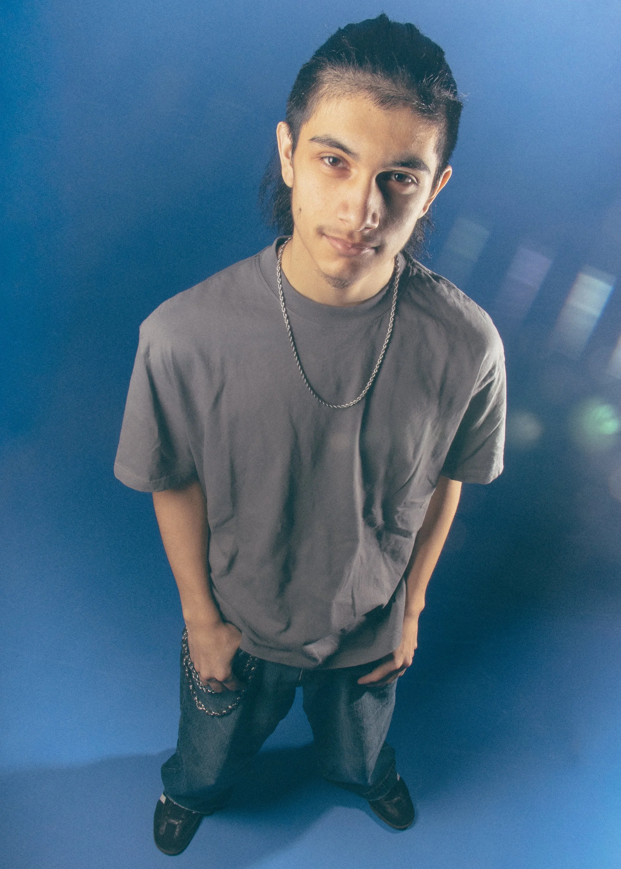 Young man with dark hair styled back, wearing a gray T-shirt, chain necklace, and jeans, standing against a blue background, looking at the camera with a slight smile.