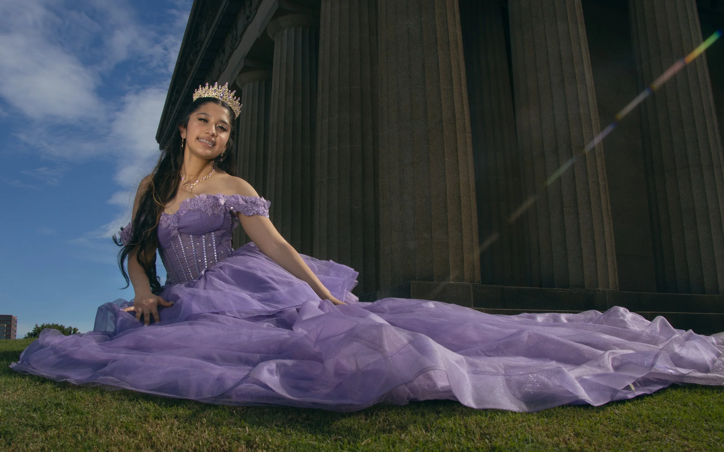 Woman in a purple gown and tiara sitting outdoors near a building with large columns, smiling at the camera.