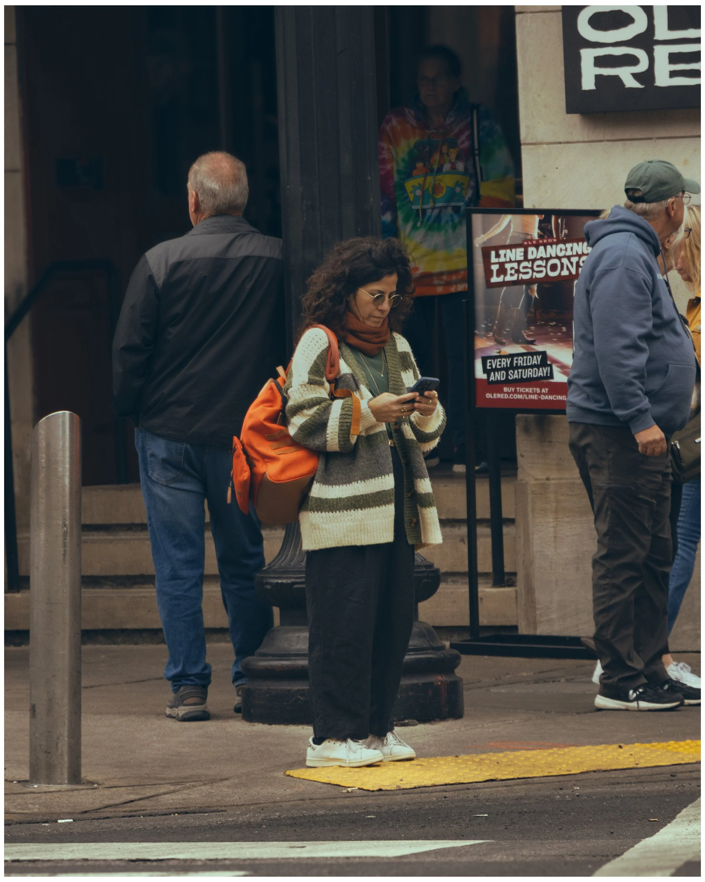 People standing at a street corner, one woman looking at her phone, others waiting, with a sign advertising line dancing lessons in the background.