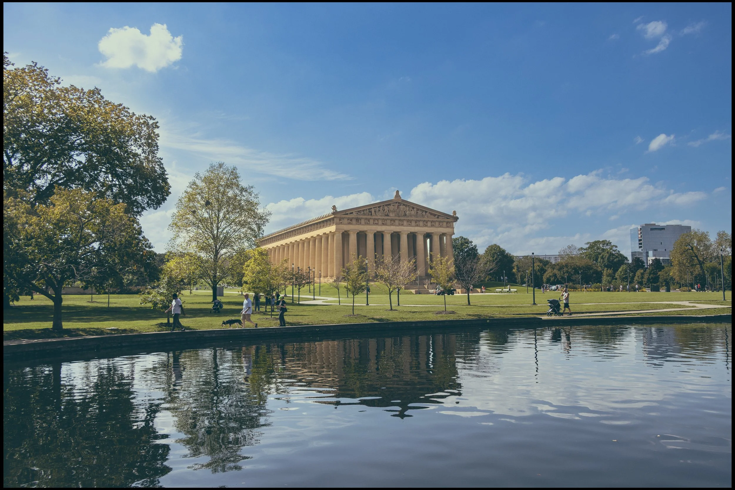 The Lincoln Memorial in Washington, D.C., with a blue sky, scattered clouds, and a reflecting pool in the foreground. People are walking and sitting on the grassy area near the memorial, and some trees are visible around the park.