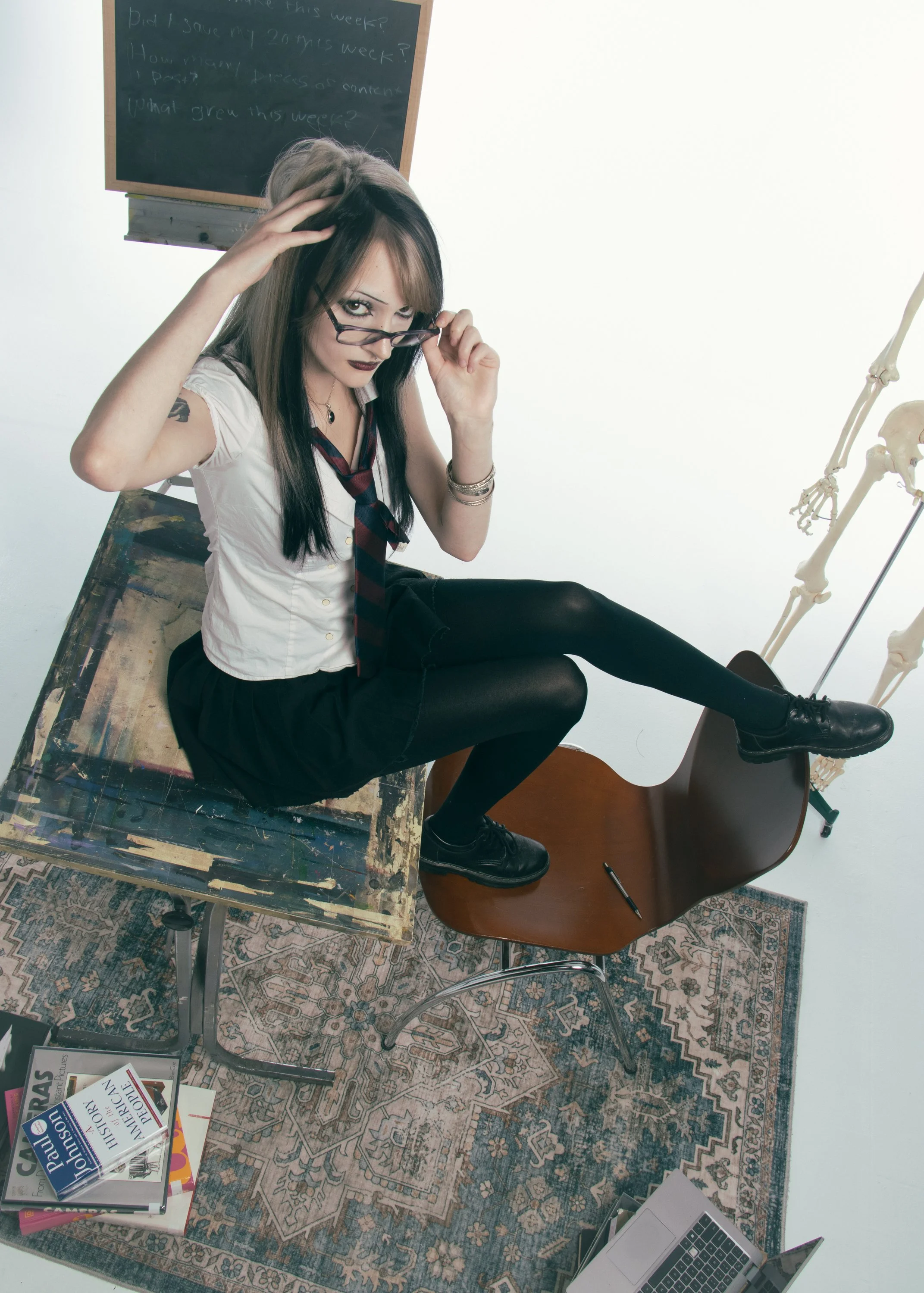 A woman with glasses, dark makeup, and fashionably styled hair, wearing a white blouse and tie, sits on a wooden chair with a skateboard inside a room. She is adjusting her glasses and looking at the camera. Behind her is a blackboard with handwritte