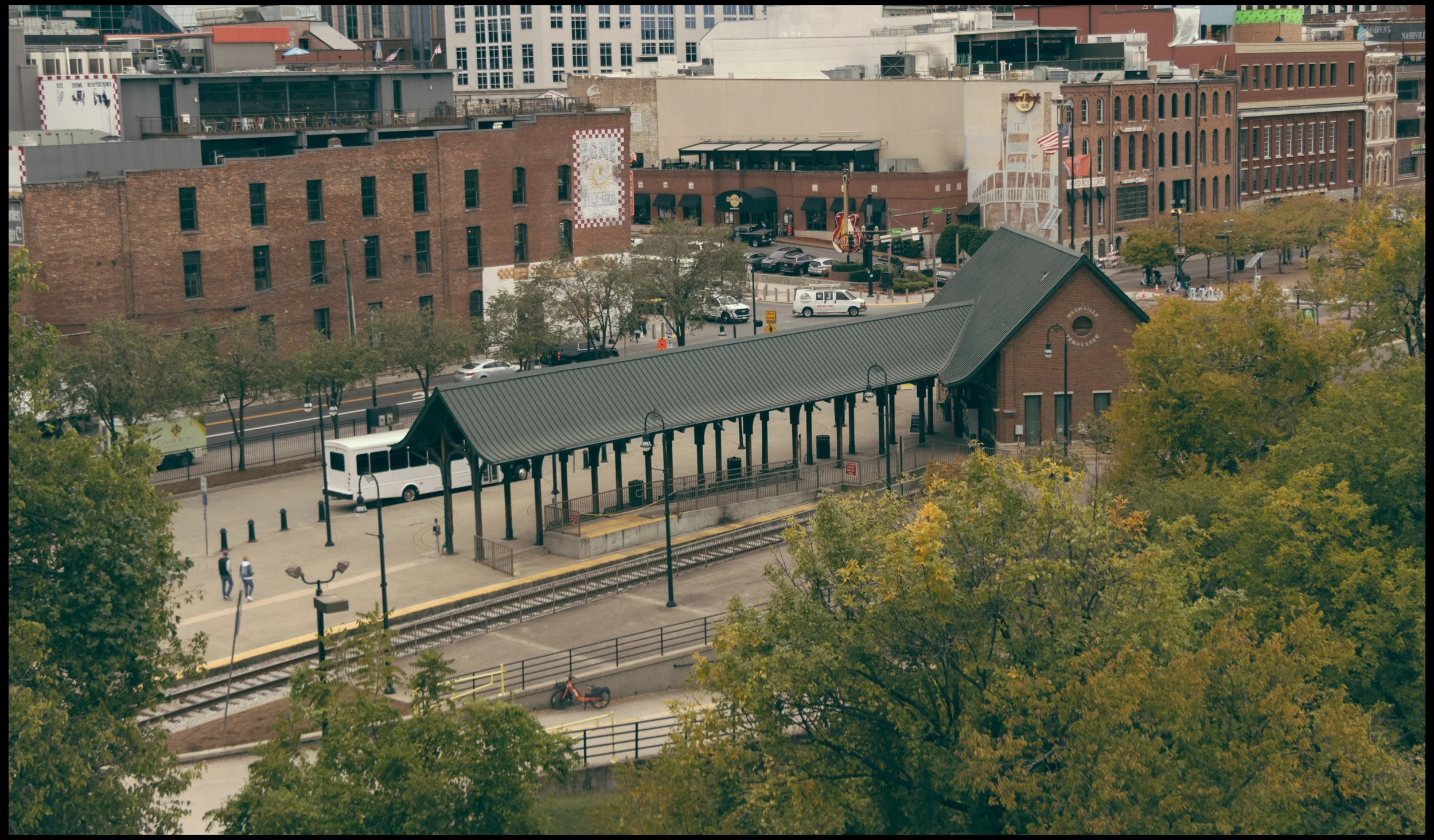 Street view with a train station platform in the foreground, several pedestrians, and a cityscape of brick and modern buildings in the background. Trees with autumn foliage are visible.