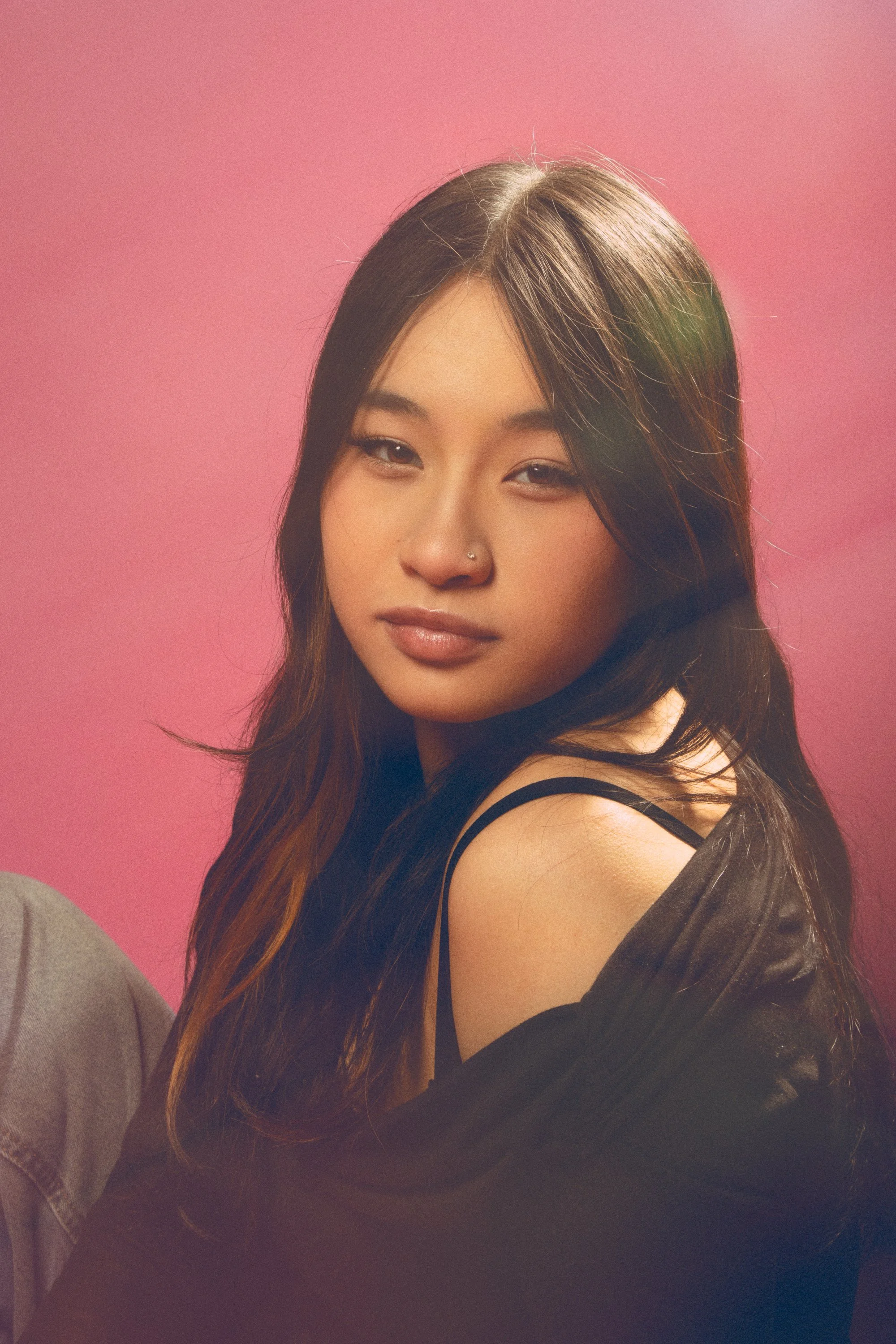 Close-up portrait of a young woman with long, wavy hair, wearing a black top and a gray jacket, against a pink background.
