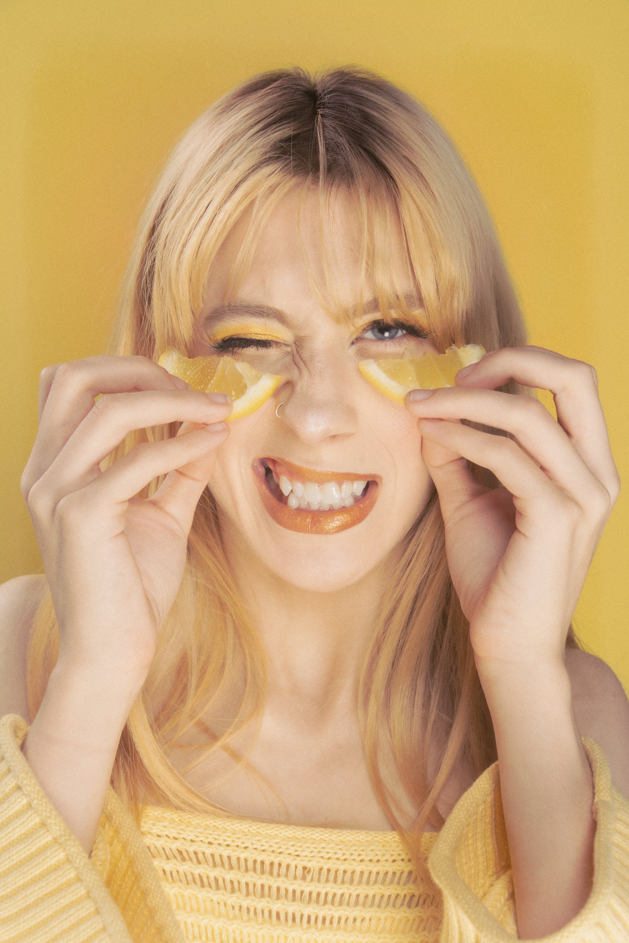 A woman with blonde hair and blue eyes holding lemon wedges over her eyes, smiling, with a bright yellow background and wearing a yellow knit sweater.