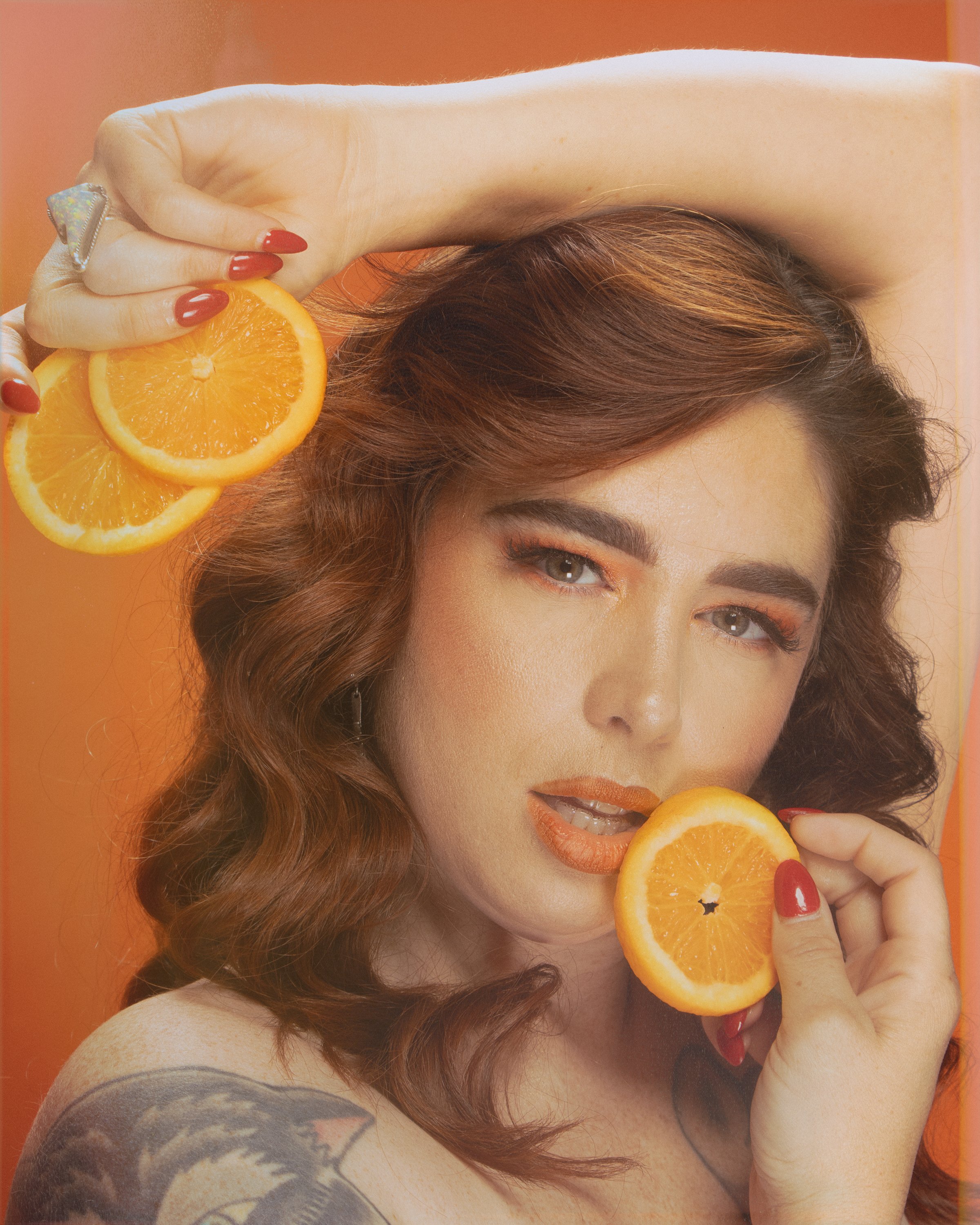 A woman with auburn hair and tattoos on her shoulder is holding a slice of orange near her mouth and another slice above her head against an orange background.