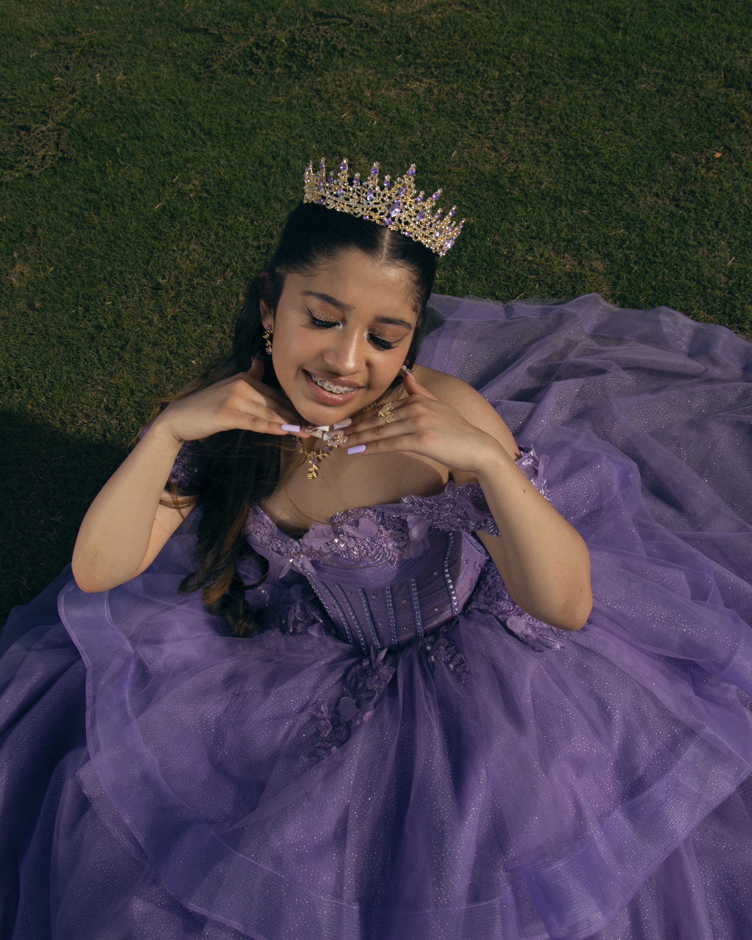 A young woman lying on the ground on a grassy field, wearing a purple dress and a sparkly tiara, smiling and resting her chin on her hands.