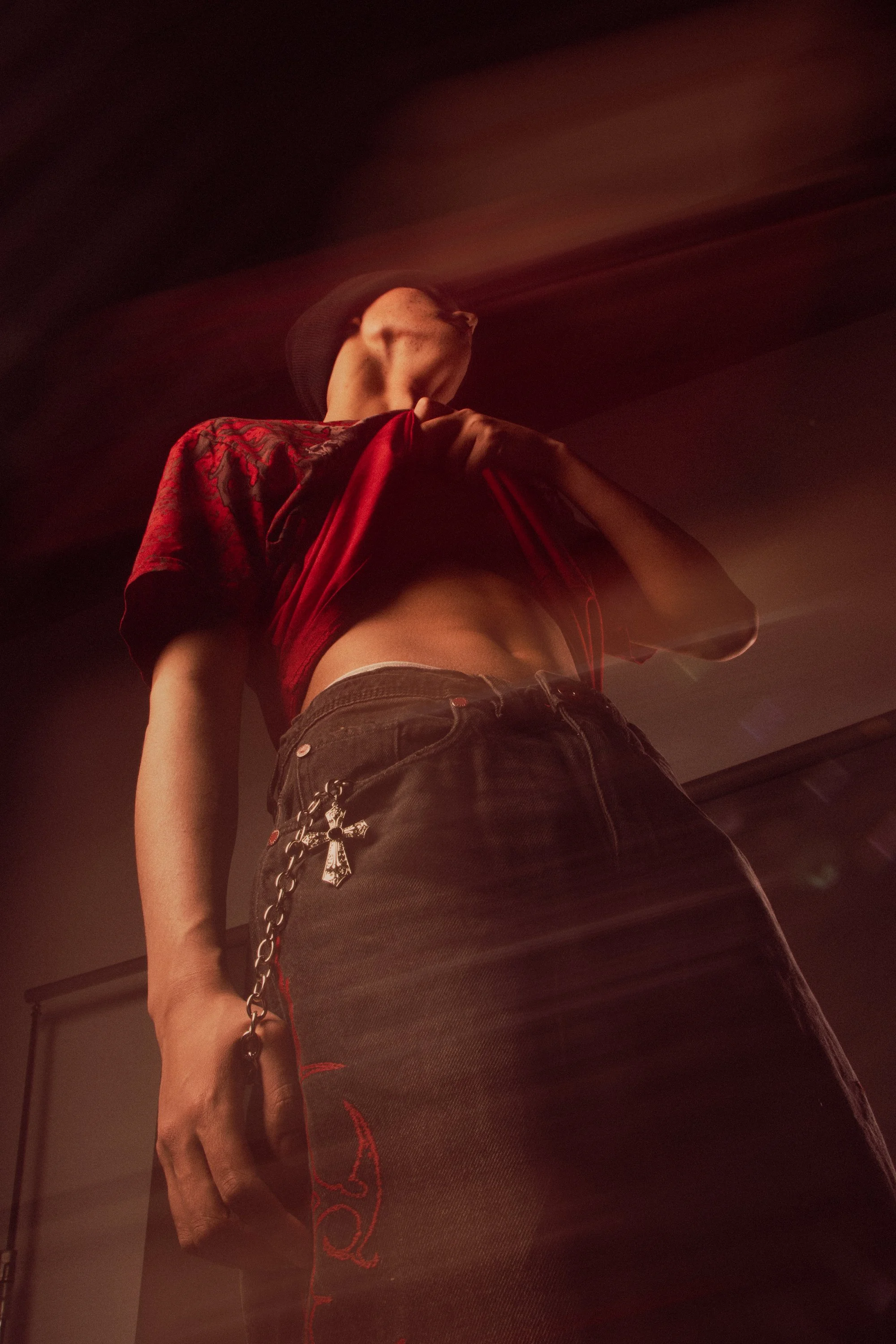 A person standing in a dimly lit room, wearing a red and black shirt, black pants, a chain with a pendant, and a black cap. The photo is taken from a low angle, showing the person's torso and part of their face.