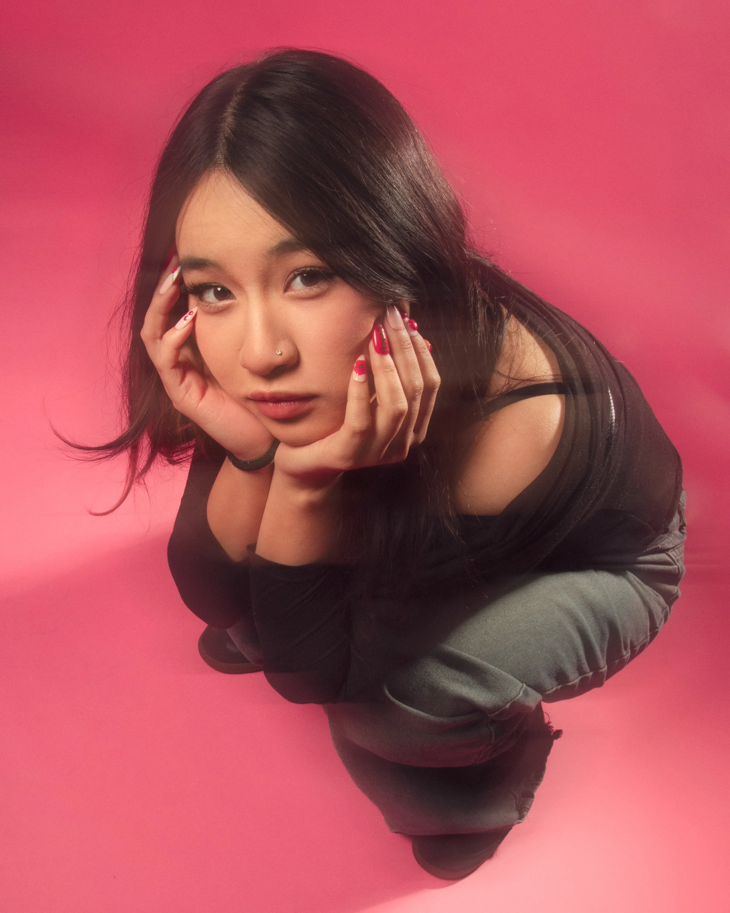 Young woman with long dark hair, wearing a black sleeveless top and gray pants, poses with her hands framing her face against a pink background.