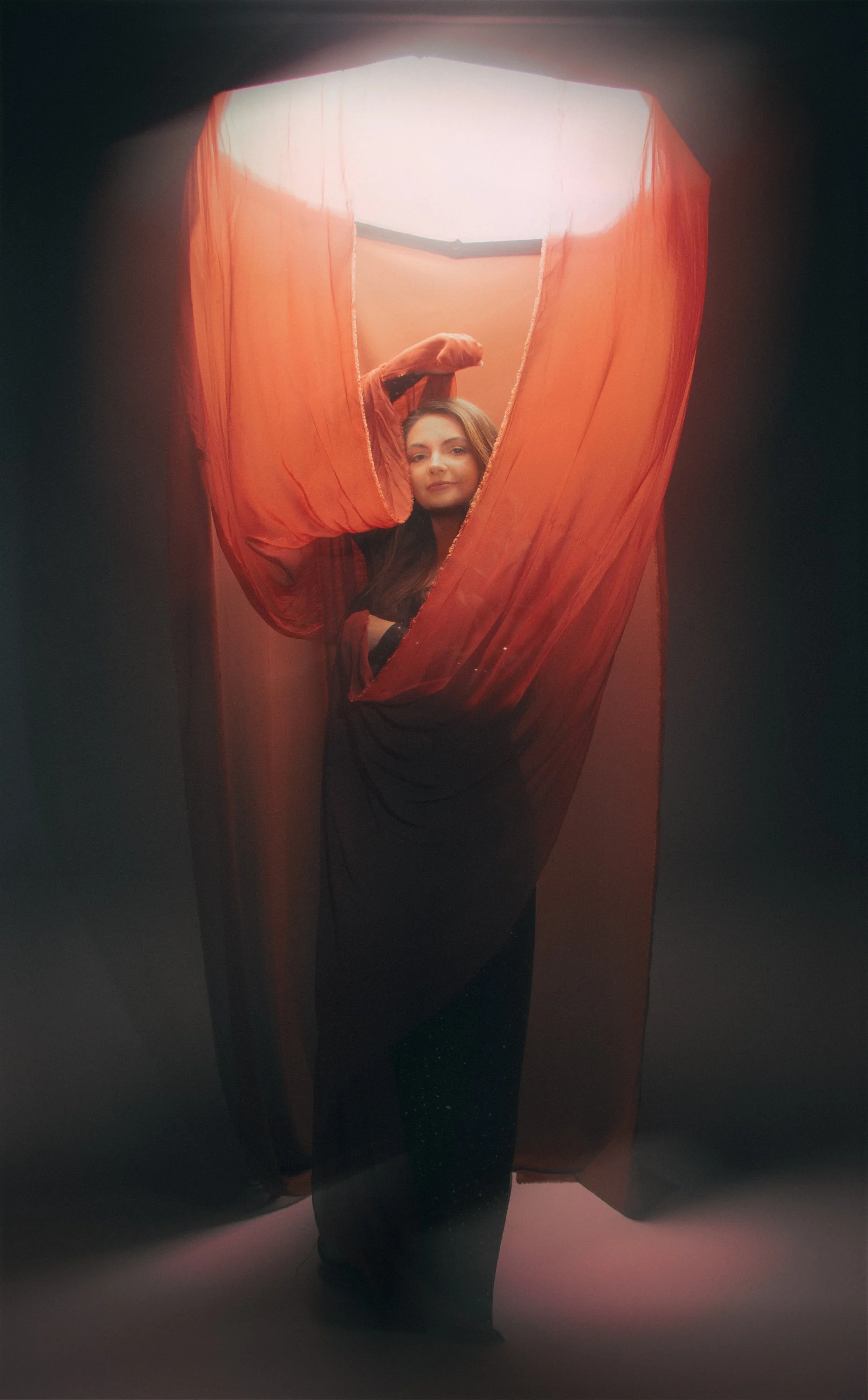 A woman standing behind a large, flowing orange fabric, with light coming from above, creating a soft, artistic glow.