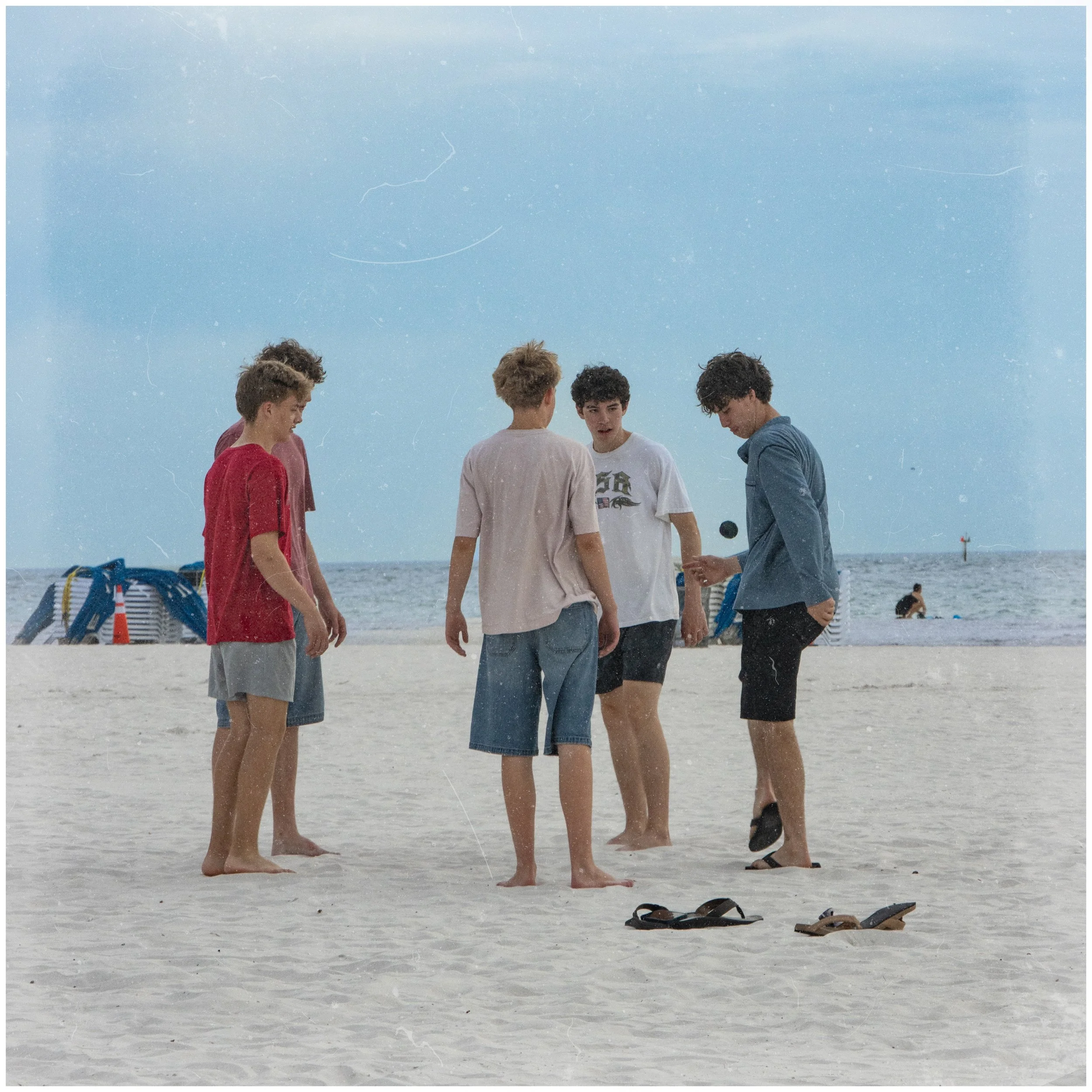 Five young men standing on a sandy beach in a circle, facing inward, with their heads bowed and engaged in conversation. The sky is partly cloudy, and there are beach chairs and a lifeguard tower in the background.