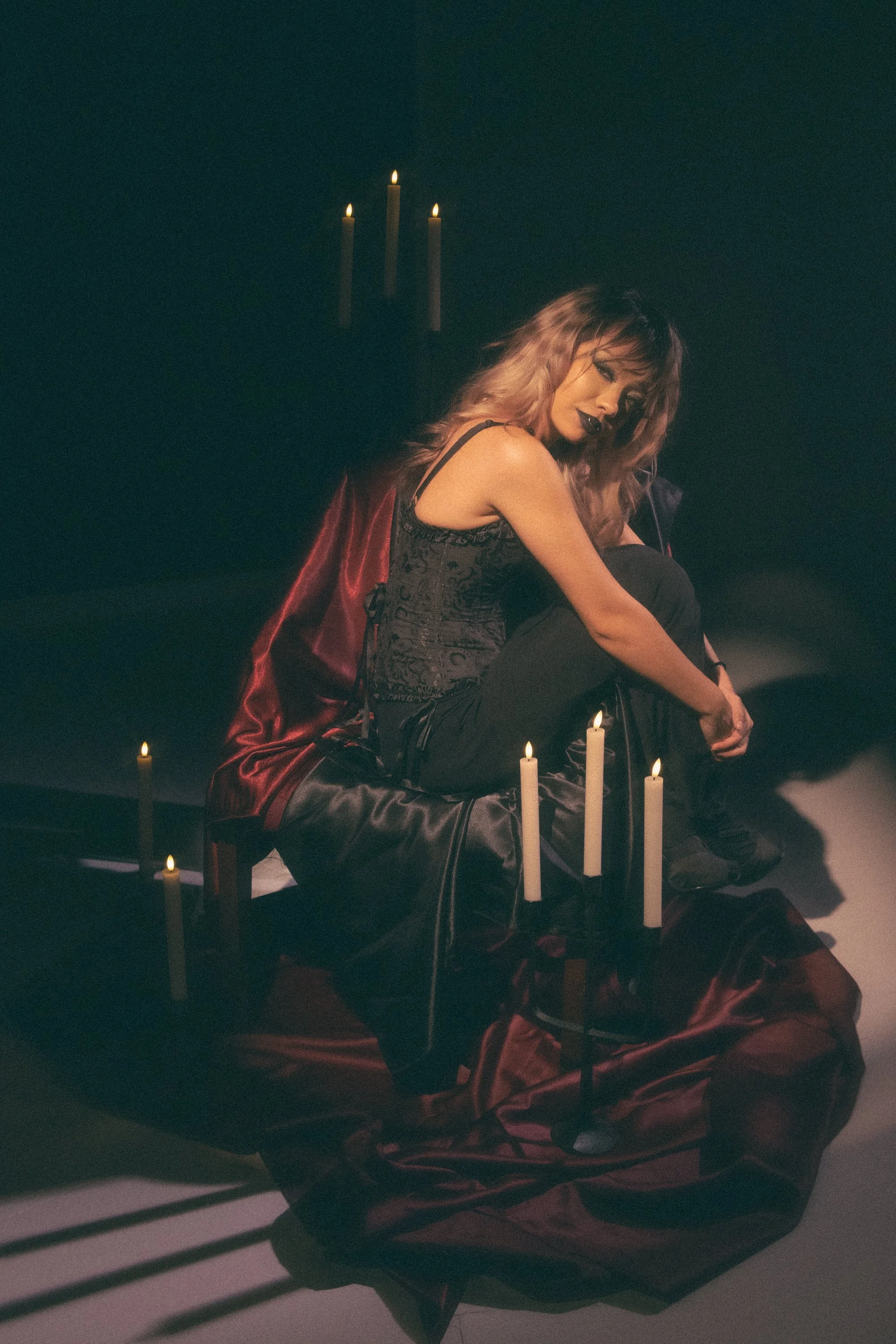 A woman with pinkish hair sitting on a black chair surrounded by lit candles with a black background and a three-candle candelabrum behind her.