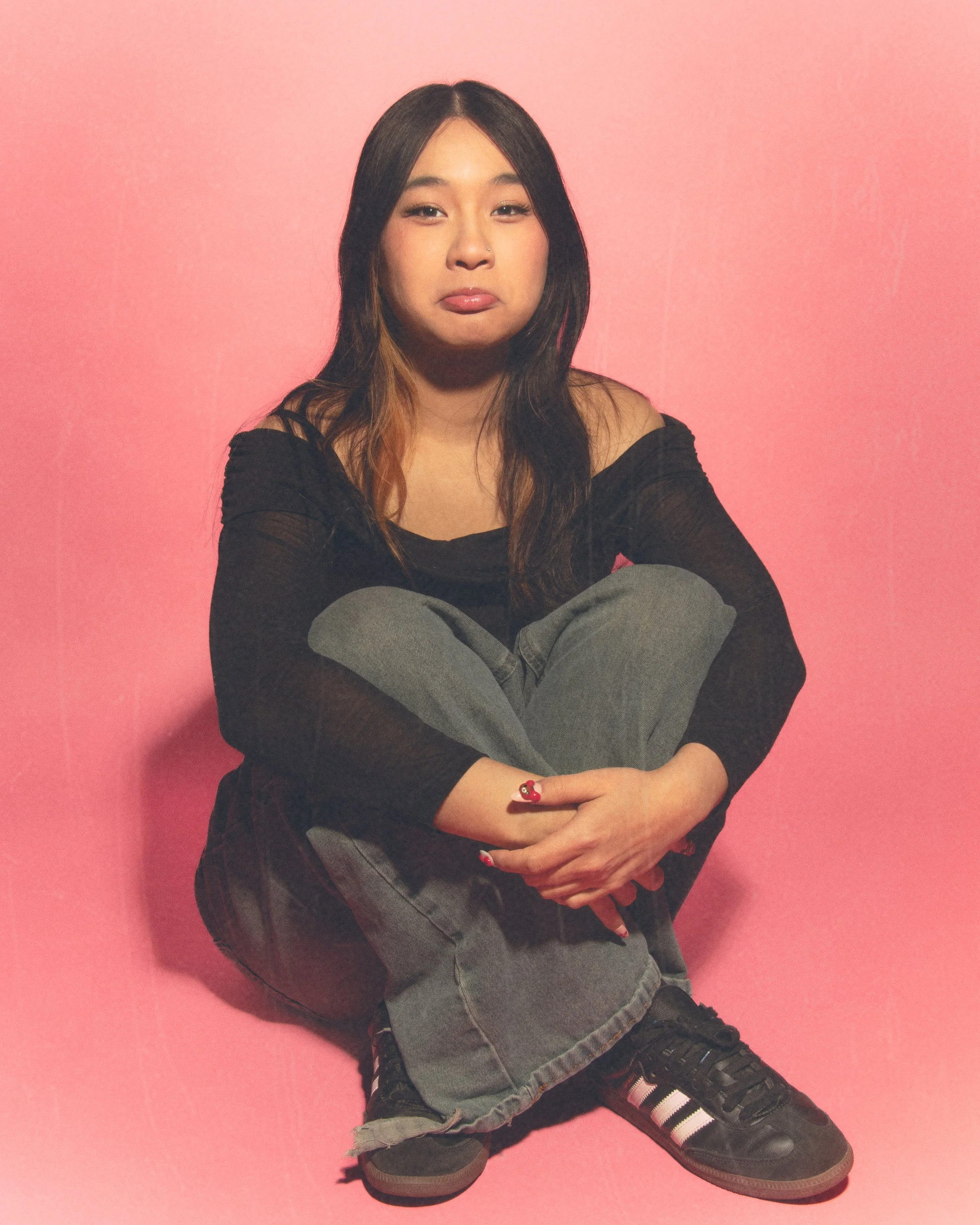 A young woman sitting on a pink floor against a pink wall with crossed arms, wearing a black off-shoulder top, gray pants, and black sneakers with white stripes.