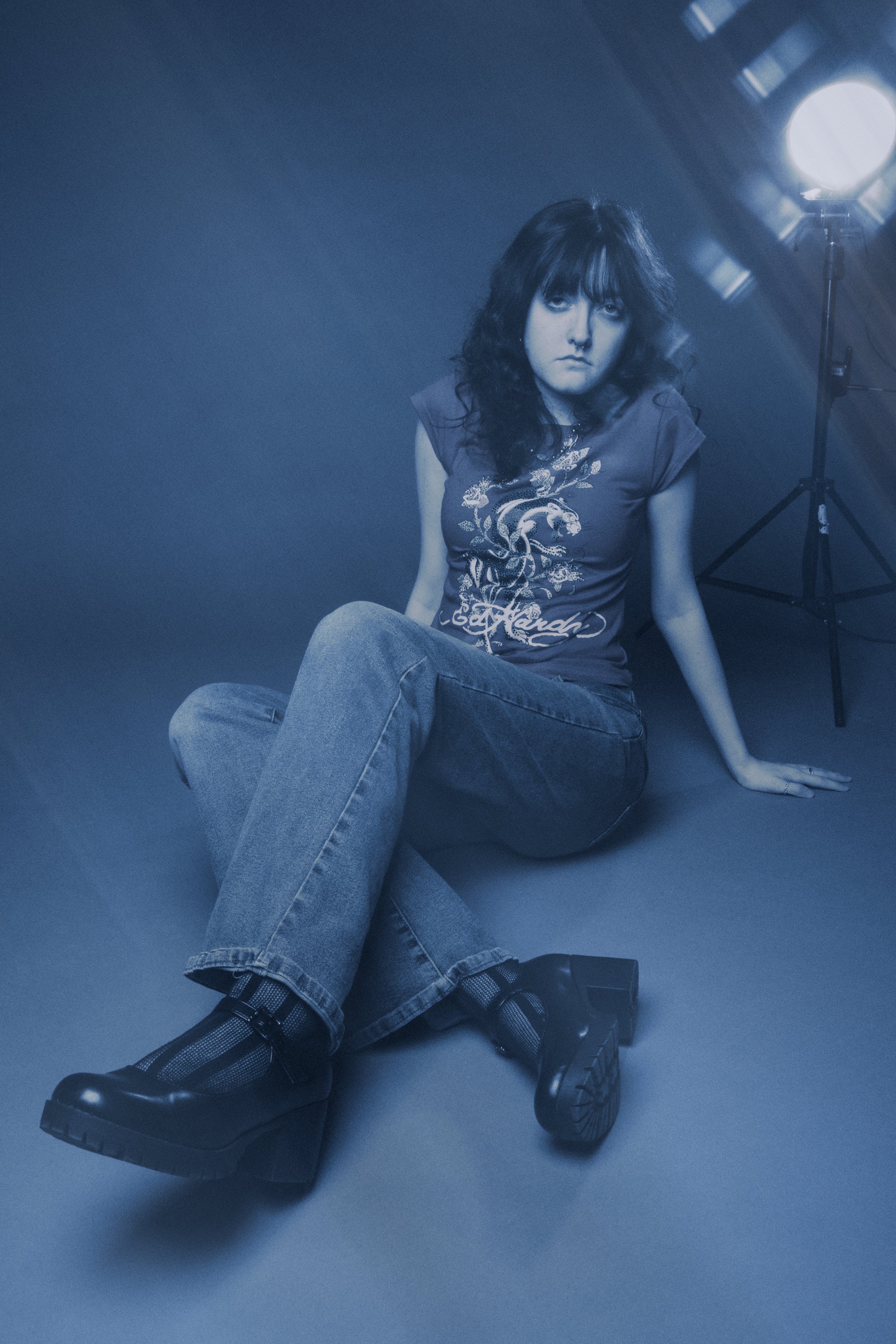 A person with dark, slightly curly hair sits on the floor with their legs crossed, wearing a t-shirt with a dragon design, jeans, and platform boots. They are in a studio with a light source and a dark background.