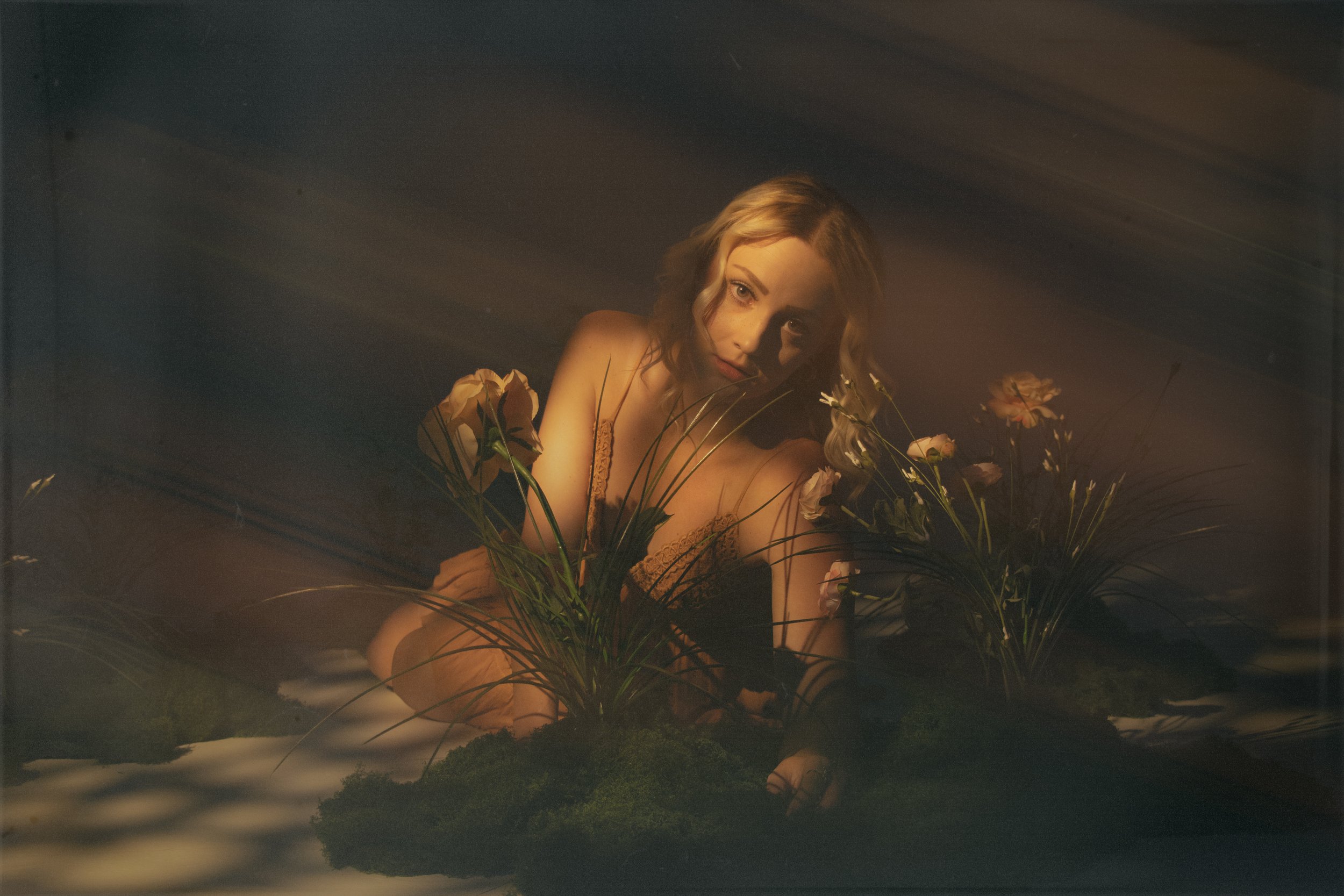 A woman with blonde wavy hair and makeup, lying among pink and orange flowers, gazing at the camera, with a soft, warm lighting effect.