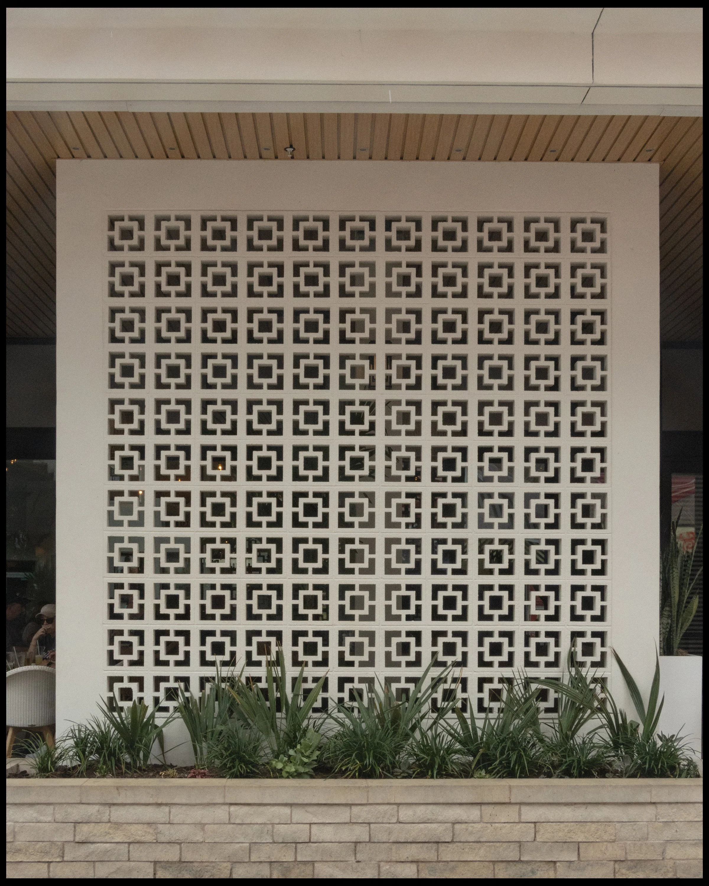 A decorative white concrete block wall with a geometric pattern, located above a brick planter filled with green plants, in front of a building with a wooden ceiling and beige wall.
