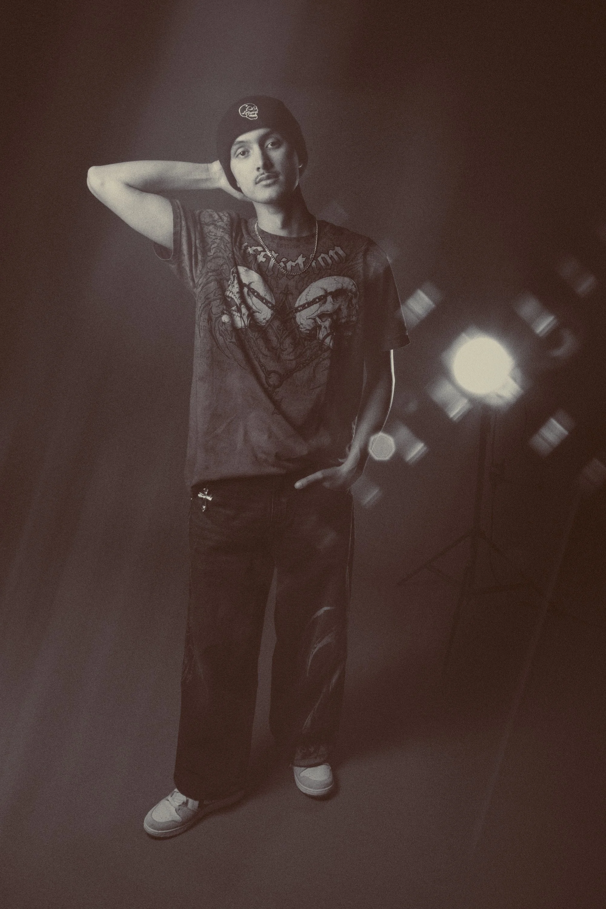A young man wearing a black beanie, a graphic t-shirt, black pants, and white sneakers, standing with one hand on his hip and the other behind his head, in front of a dark background with fluorescent lights.