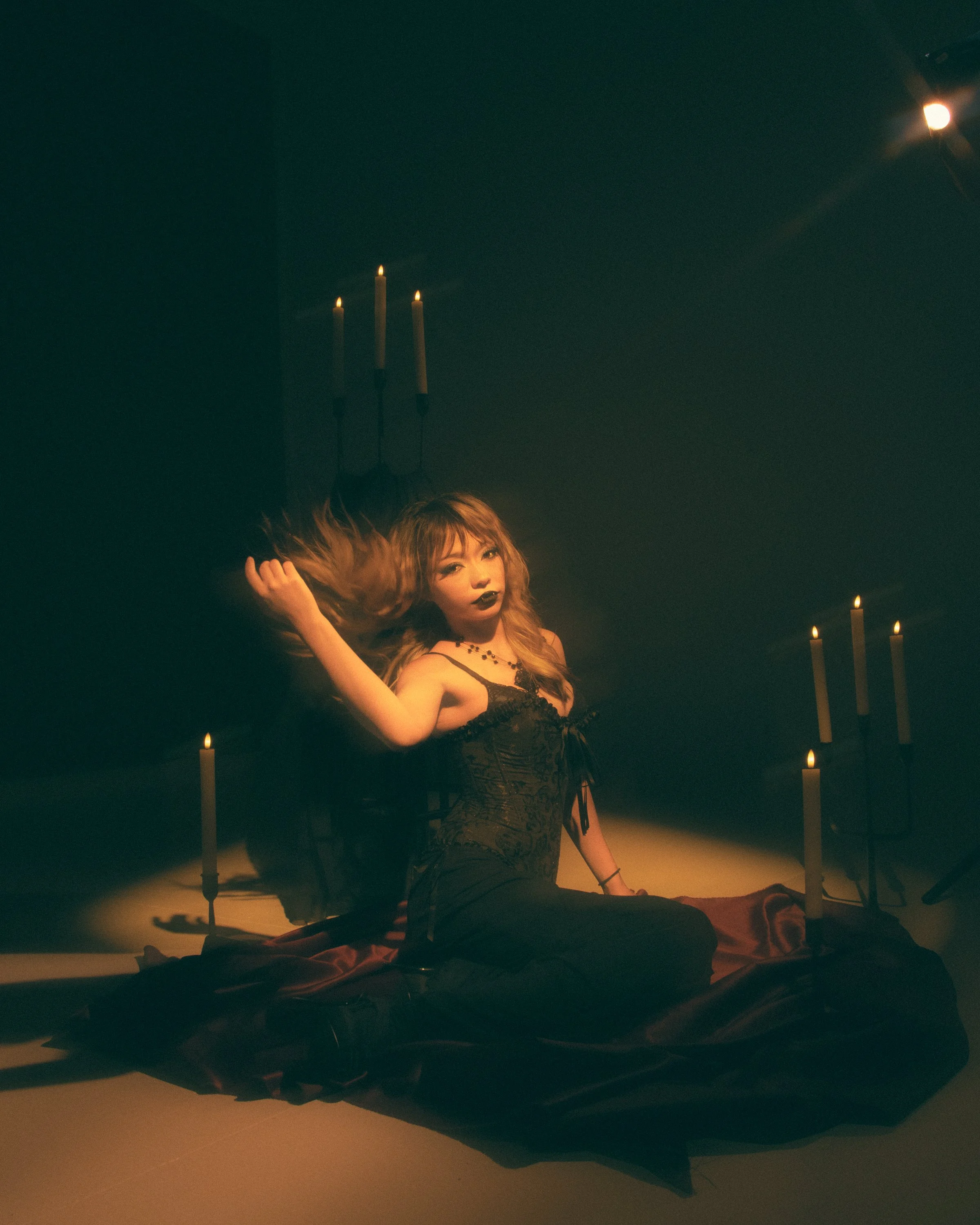 A woman with curly hair and dark makeup sitting on the floor in a dimly lit room, surrounded by black fabric and lit candles, creating a gothic or alternative aesthetic.