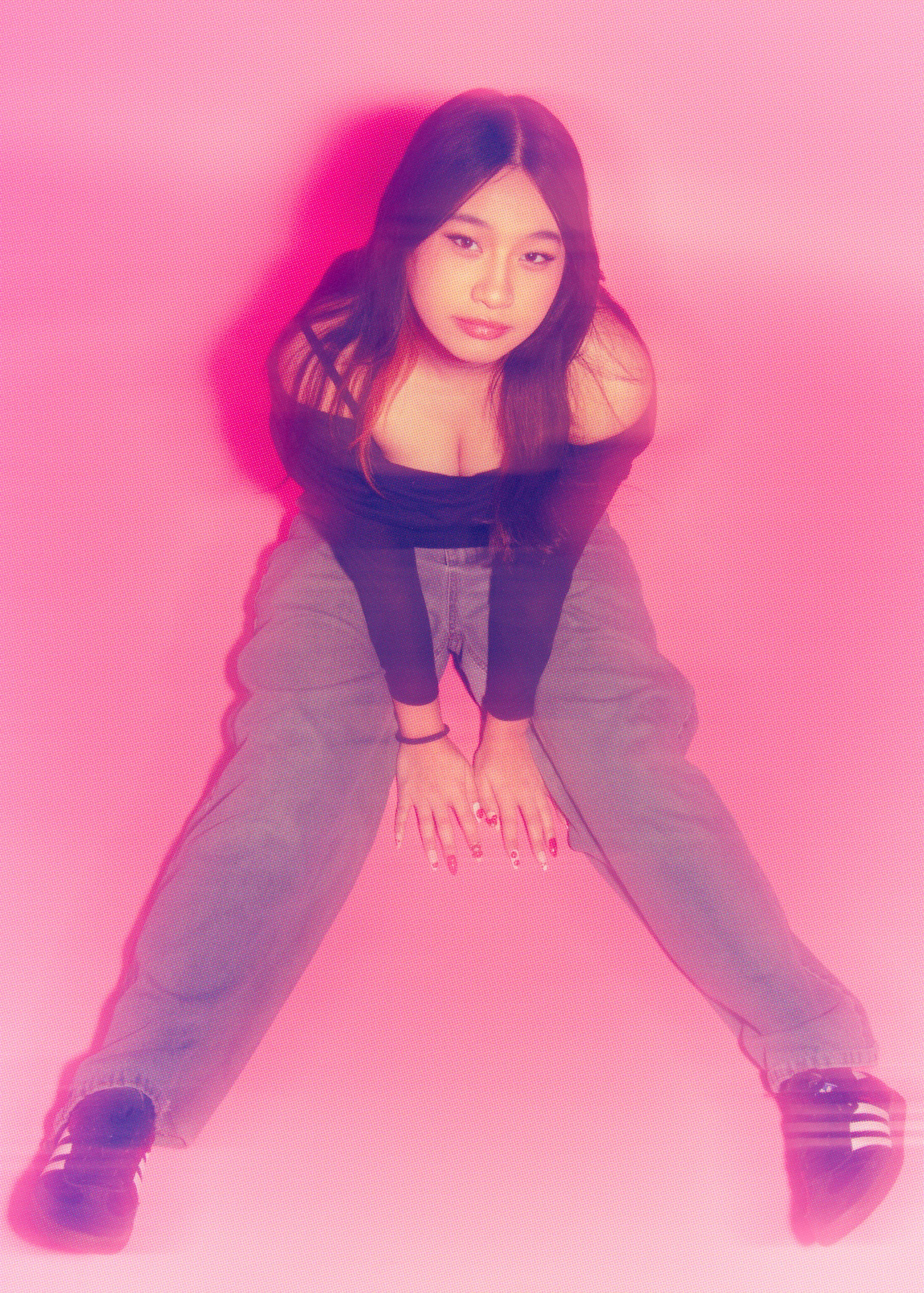 A woman with long dark hair and minimal makeup, wearing a black off-shoulder top and gray pants, is squatting with her legs apart on a pink background. She is looking up at the camera with one hand on her knee and the other on the floor, and she is w