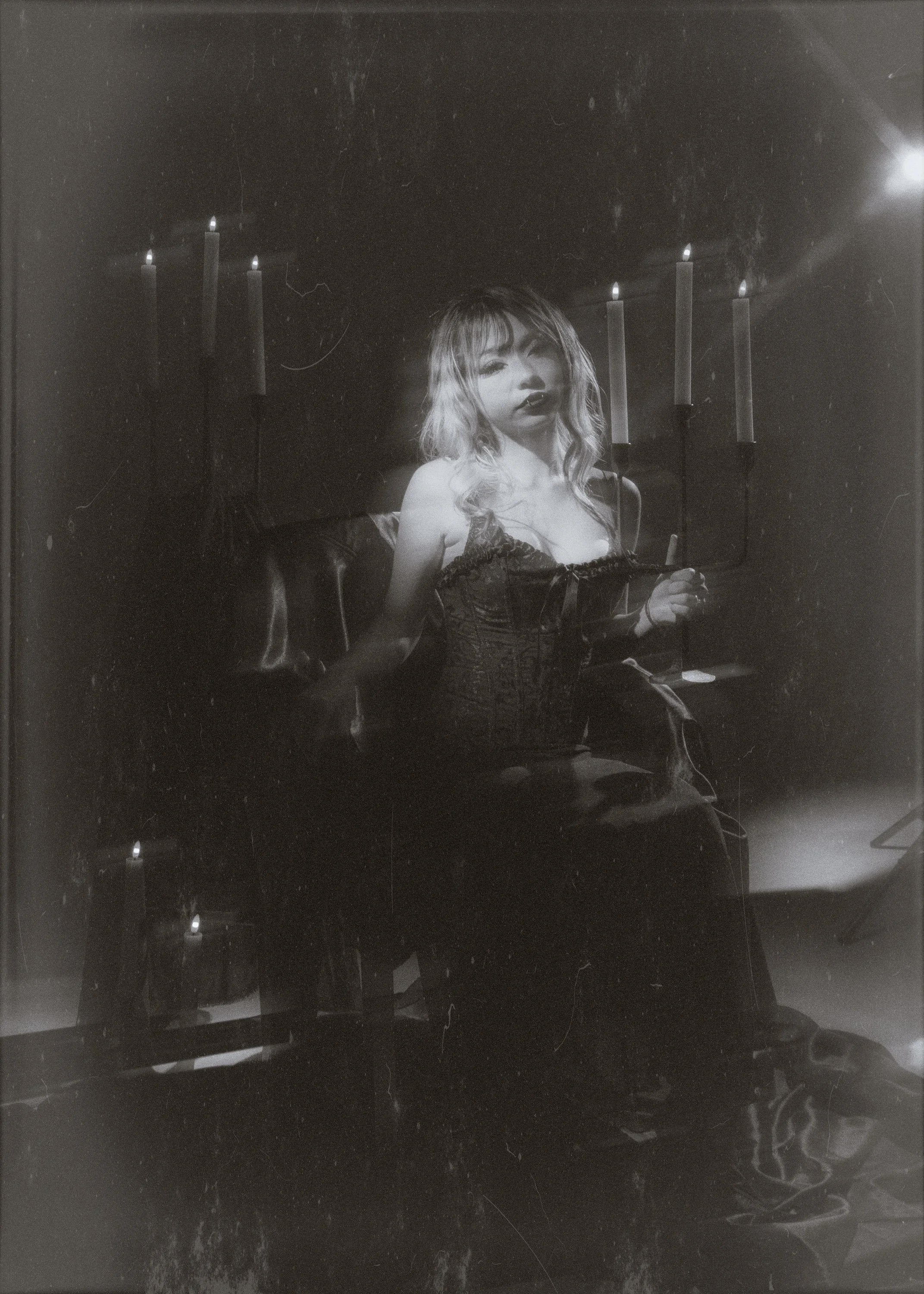 A woman with wavy blonde hair is sitting on a dark couch in a dimly lit room with candlelight and candles on the walls behind her, holding a lit candle in her hand.