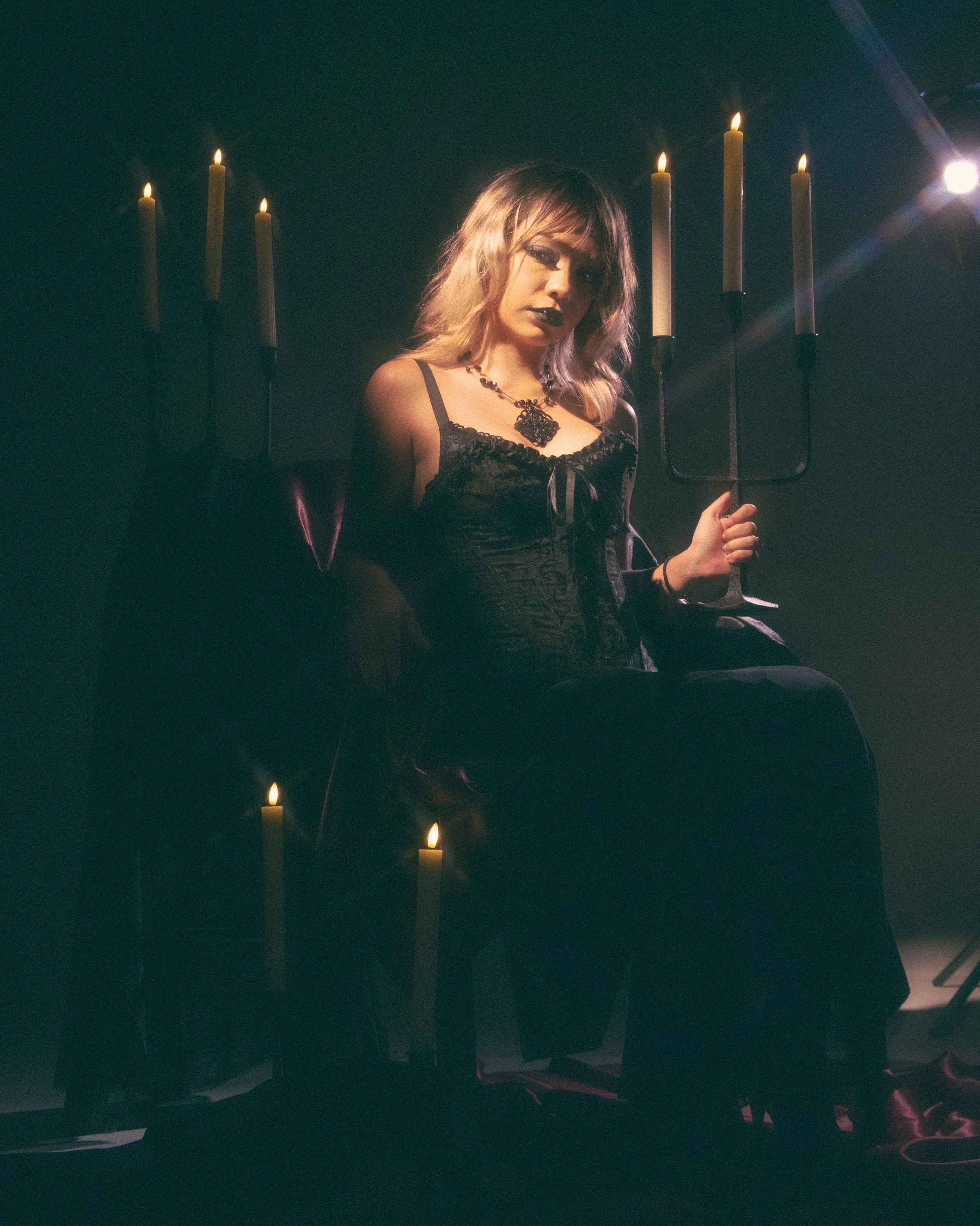 A woman with blonde hair and dark makeup, wearing a black lace dress and a black necklace, sitting in dim lighting with seven lit candles behind her, holding a black candelabrum.