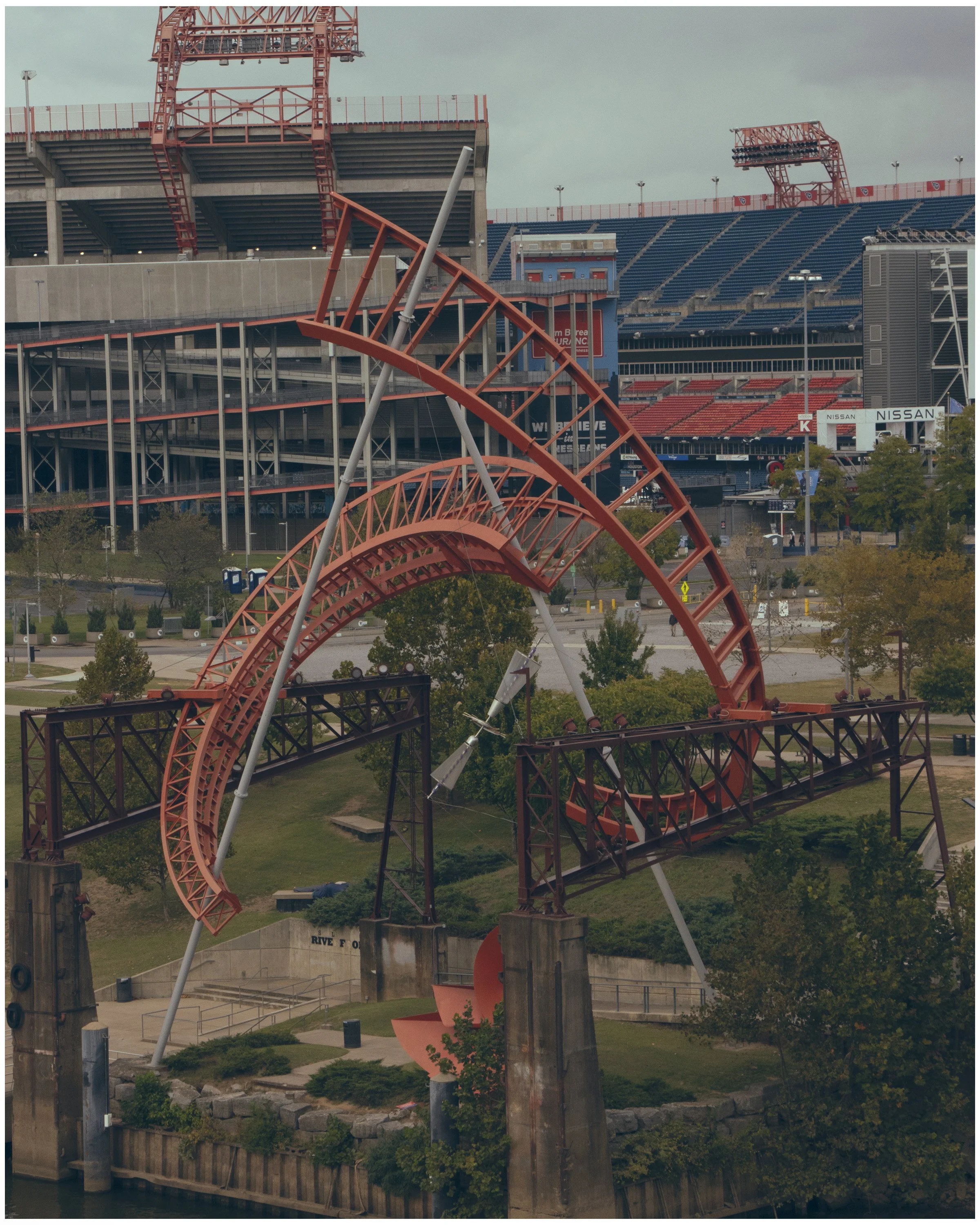 An abandoned amusement park with a damaged red roller coaster track, a river visible at the bottom, and stadium seating in the background.