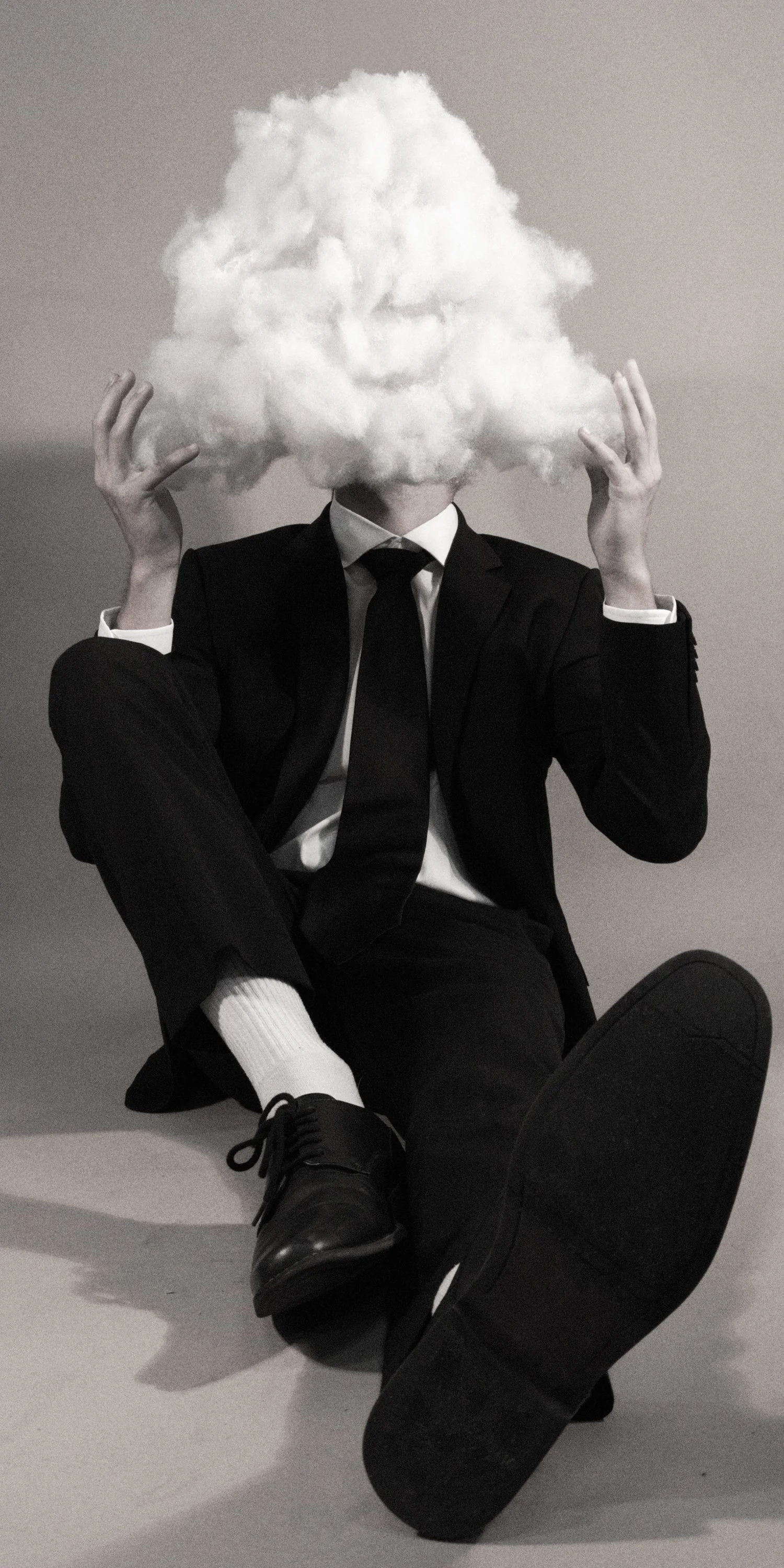 A person in a black suit, white shirt, and black tie with a cloud of smoke in the shape of a face covering their head, sitting on the floor with one leg crossed over the other.