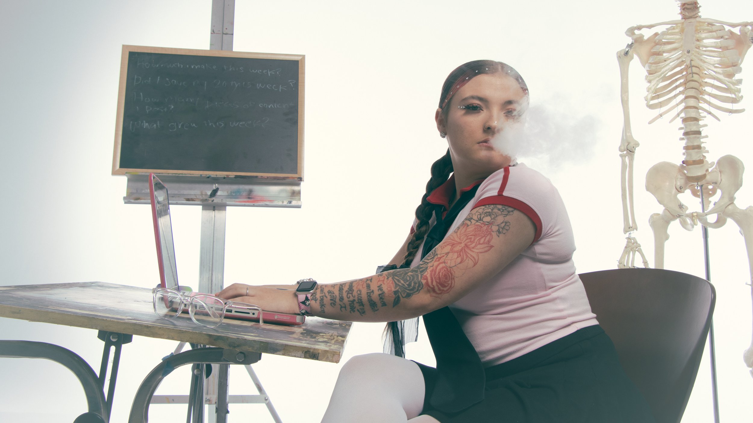 A woman with tattoos on her right arm, sitting at a desk with a pink smartphone and glasses, in a classroom setting with a skeleton model in the background and a blackboard.
