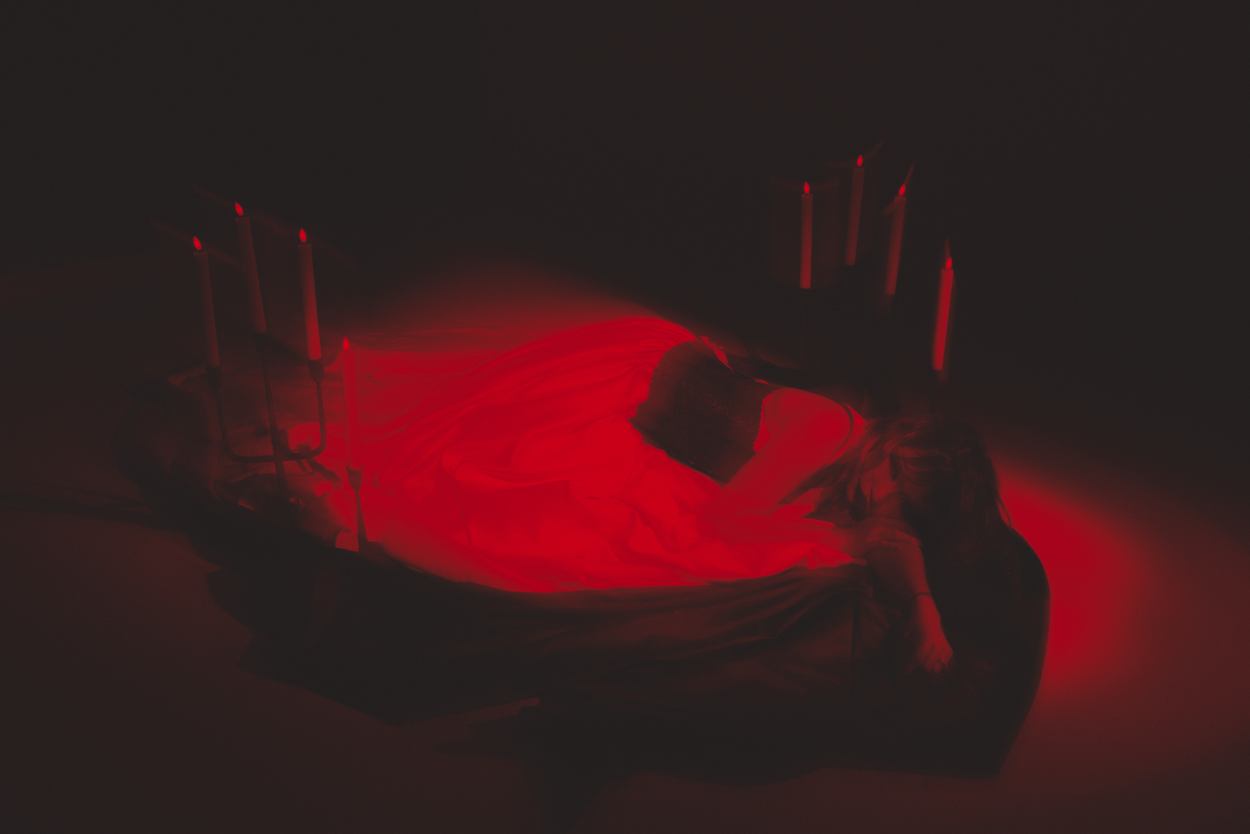 A person lying on their back with long hair, surrounded by red glowing candles, creating a dark, moody atmosphere.