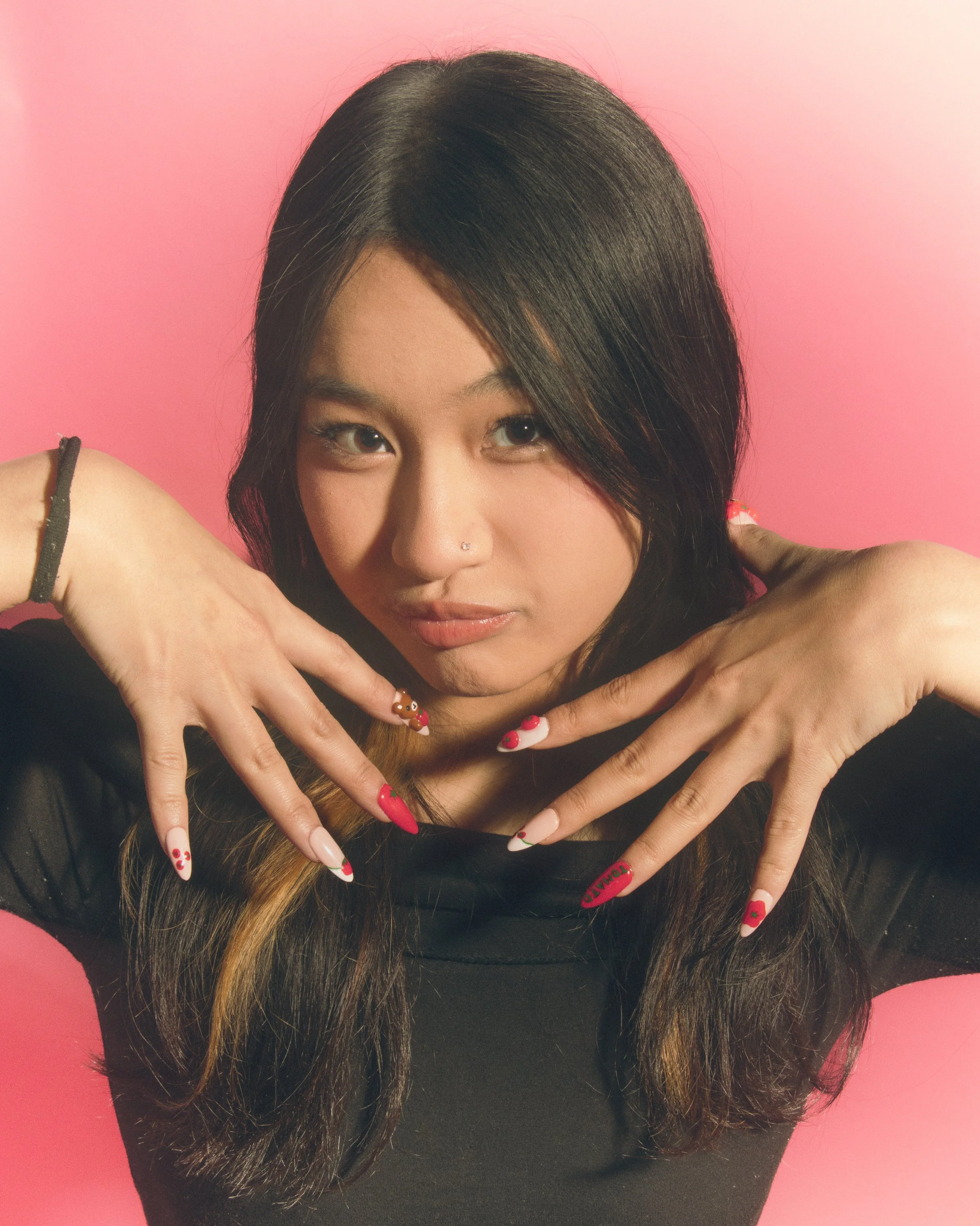 A woman with long ombre hair wearing a black top, posing with her hands near her face, against a pink background. She has a nose piercing and decorated nail art.