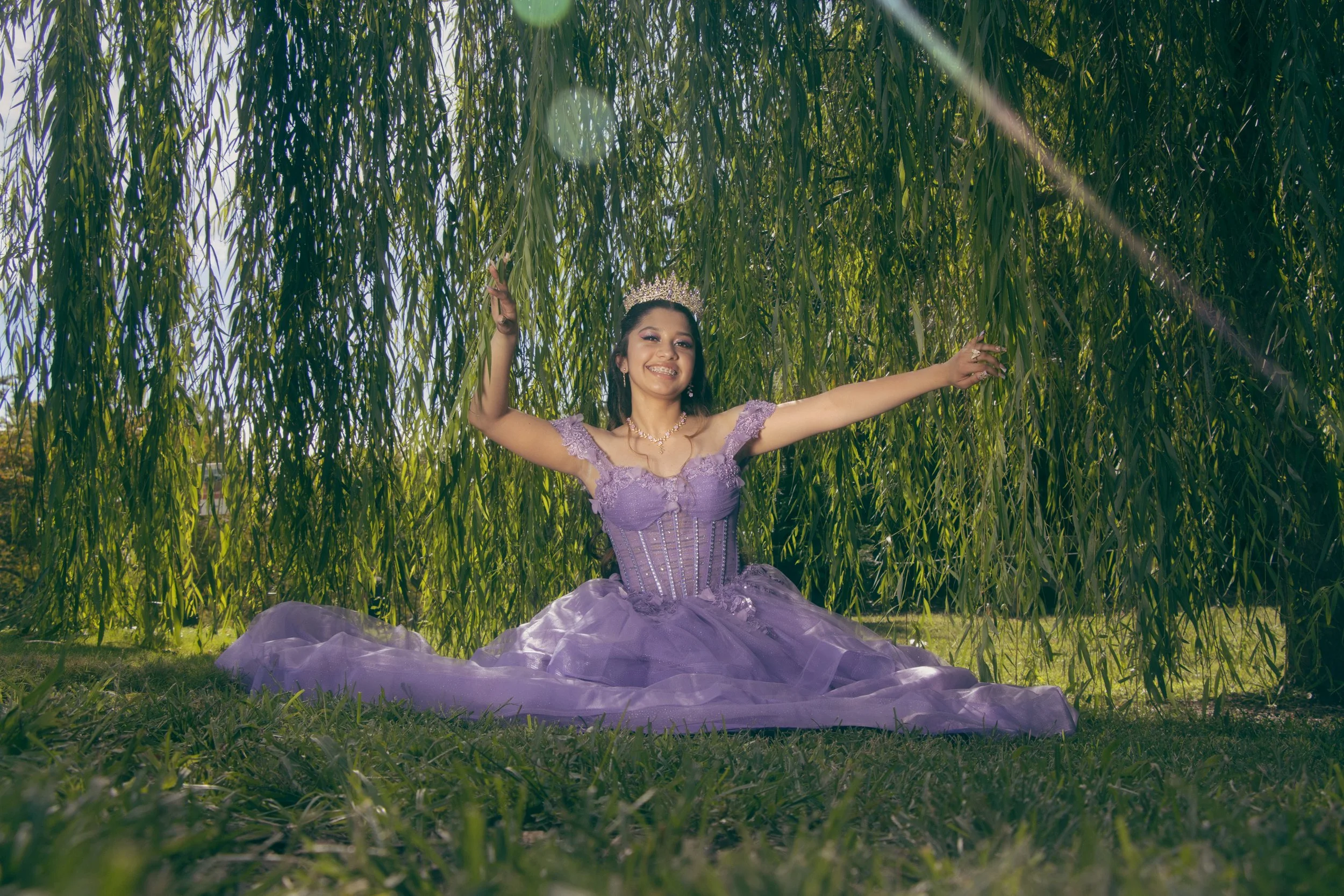 A young woman dressed as a princess in a purple gown and tiara, sitting under a tree with hanging branches, smiling and extending her arms outward.