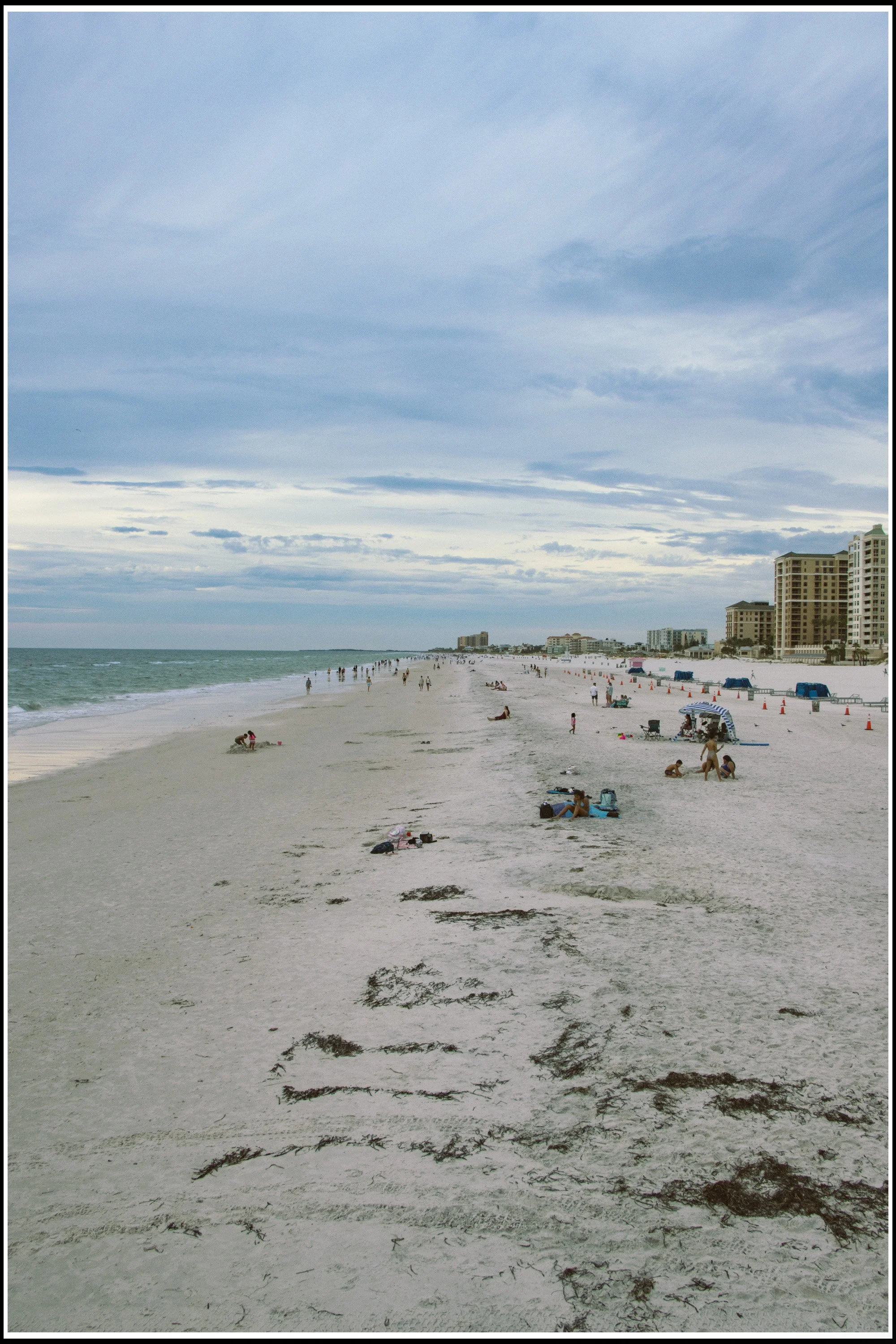 A beach scene with people relaxing and playing on the sand. The shoreline extends into the distance with ocean waves on the left. There are high-rise buildings on the right side of the image, and the sky is cloudy with scattered clouds.
