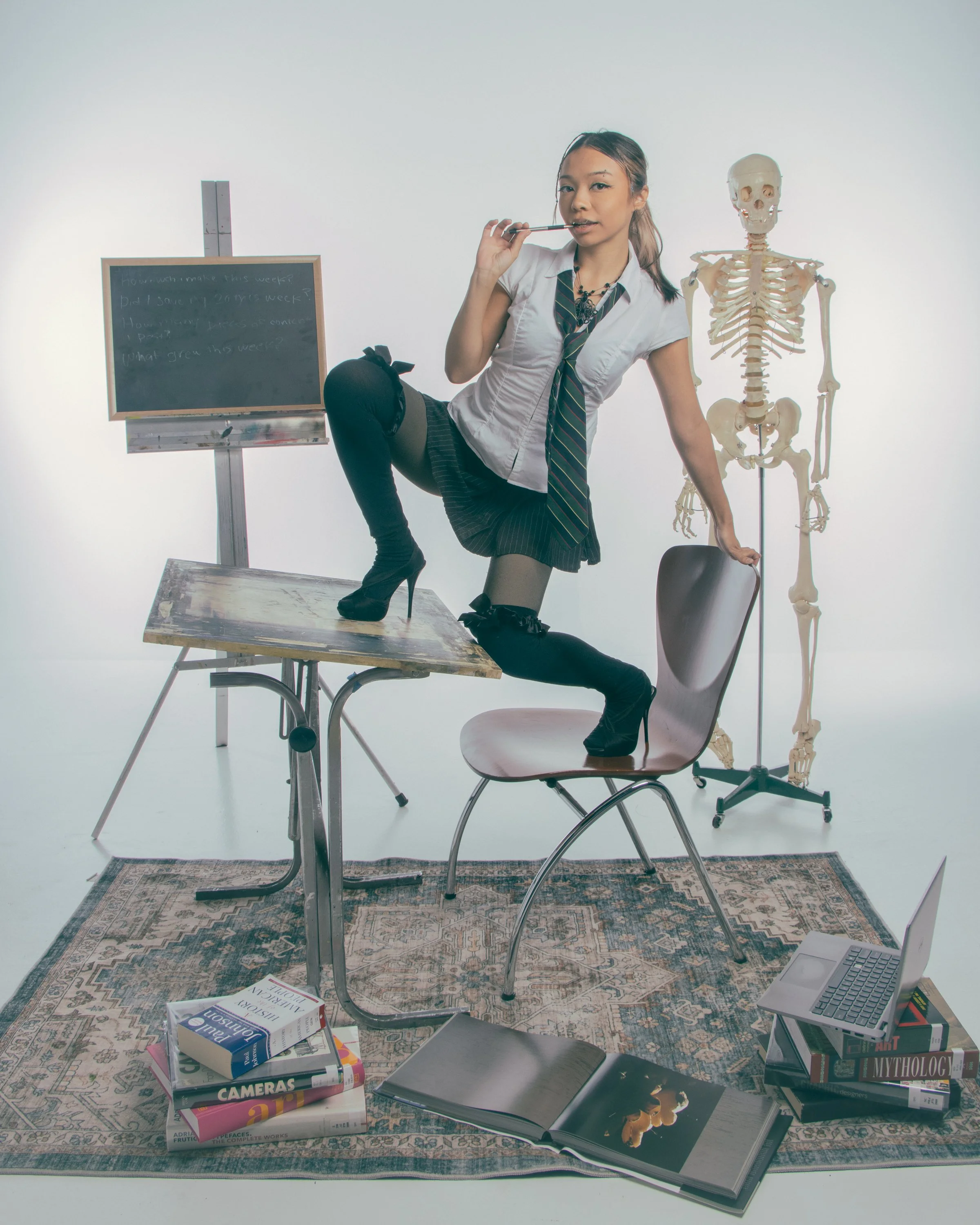 A woman in a schoolgirl outfit with thigh-high stockings and high heels poses with a skeleton in a photography studio. The studio has a blackboard with writing, a wooden table with books on photography, a laptop, and a rug.