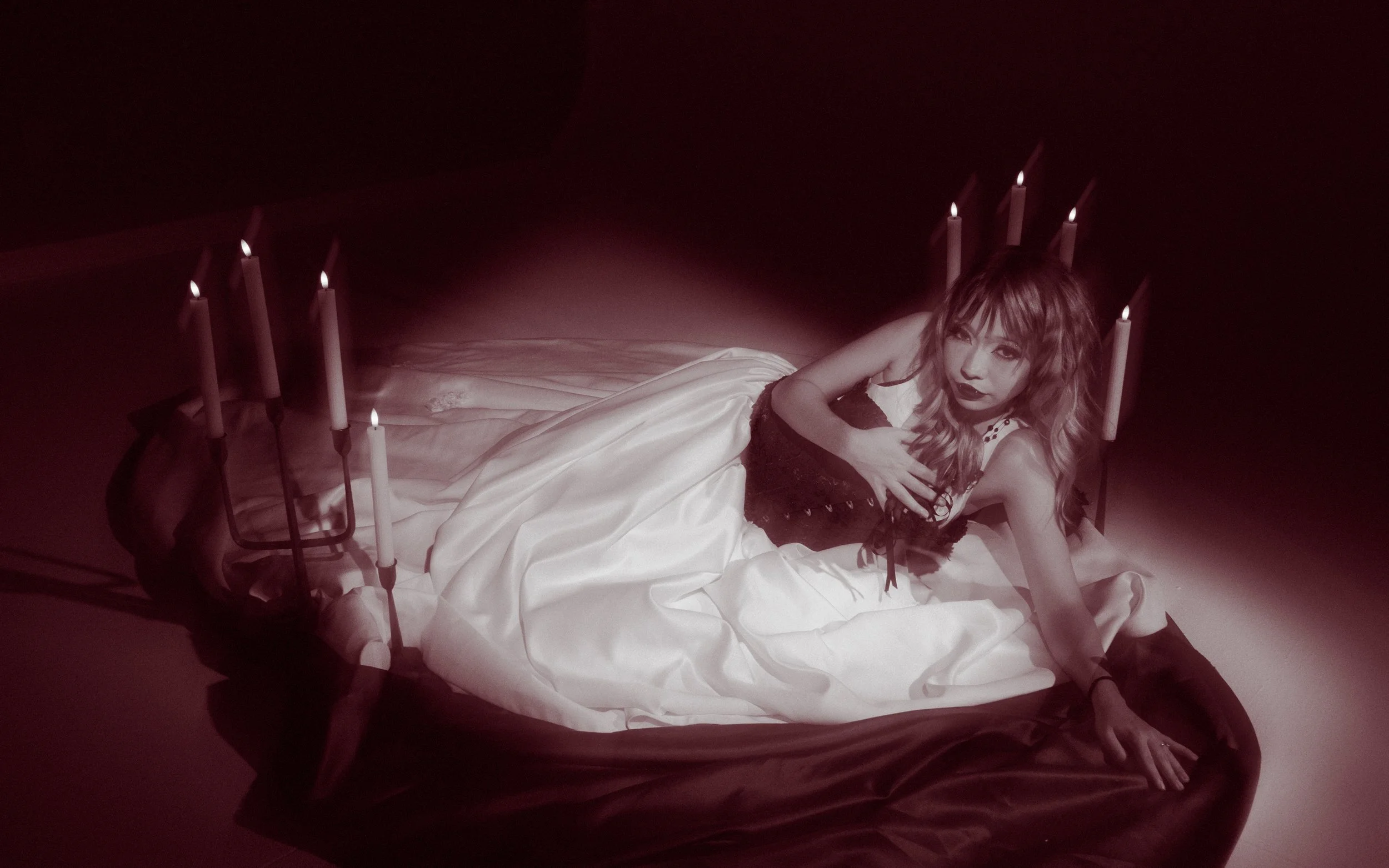 Woman lying on a bed surrounded by lit candles in a dark room.