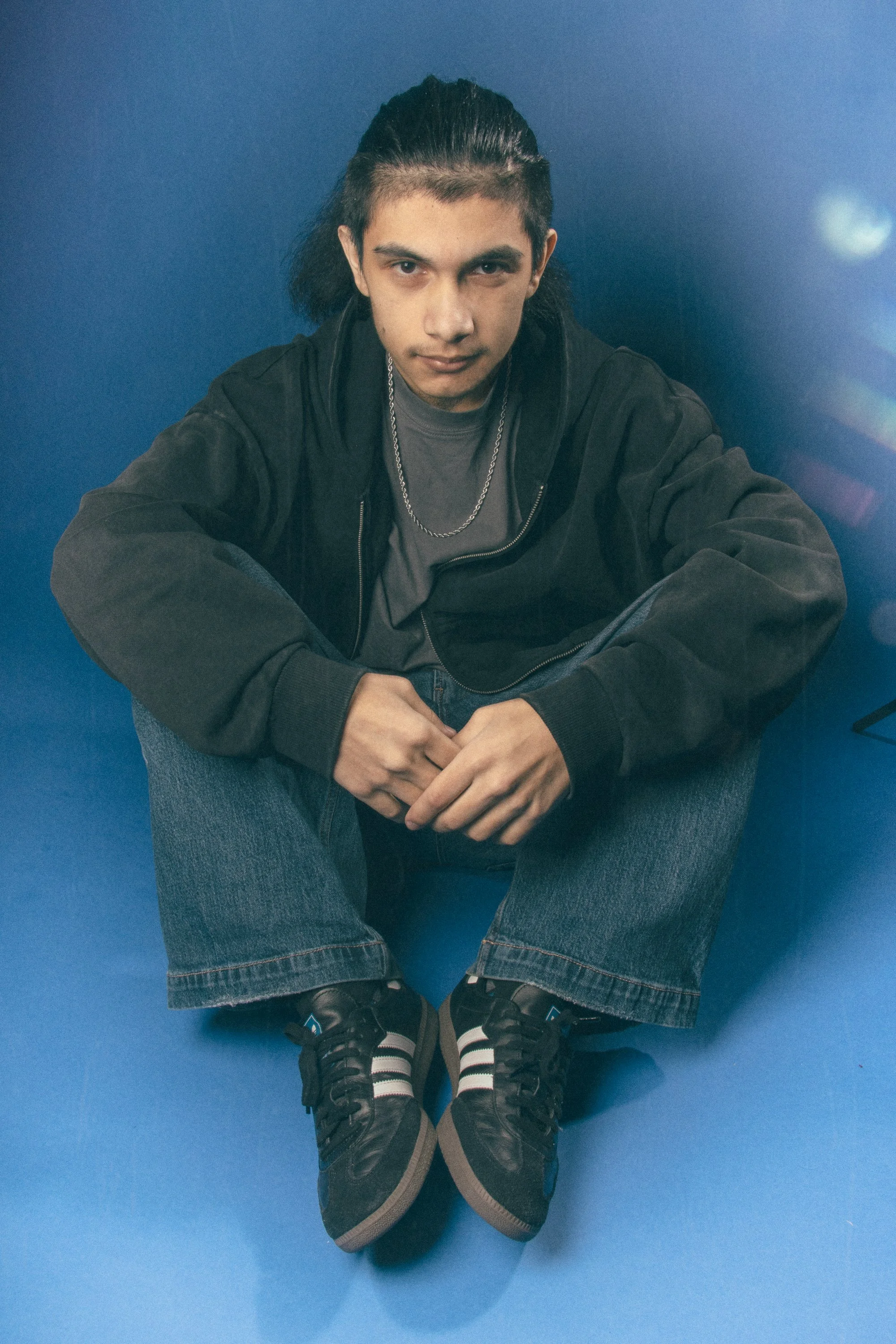 A young man with dark hair slicked back, wearing a dark jacket, gray t-shirt, jeans, and athletic shoes, sitting on the floor against a blue background. He looks directly at the camera with a confident expression.