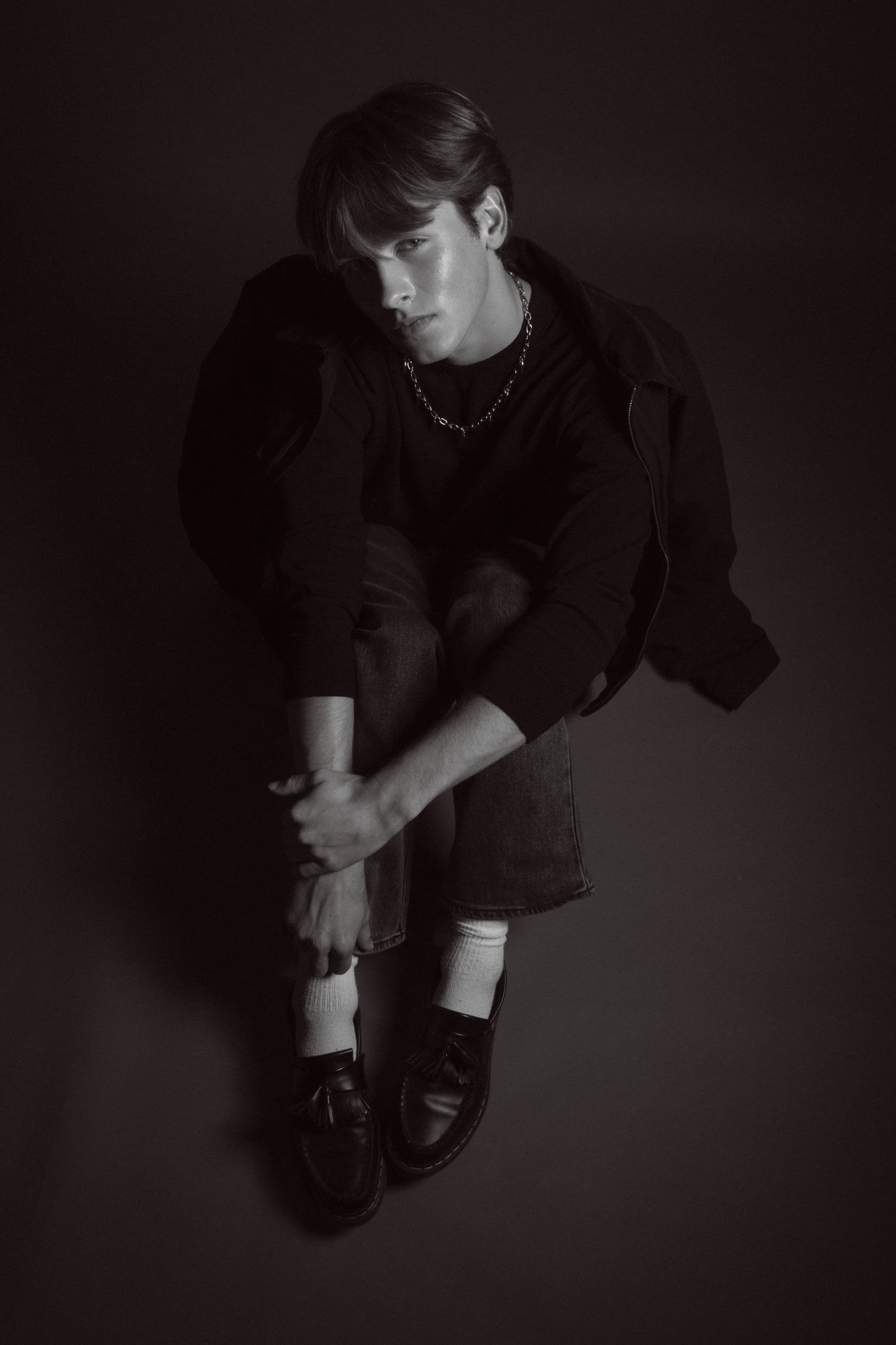 A black and white photo of a young man with styled hair, wearing a black hoodie, chain necklace, denim jeans, white socks, and black shoes, sitting with knees bent, in front of a dark background.