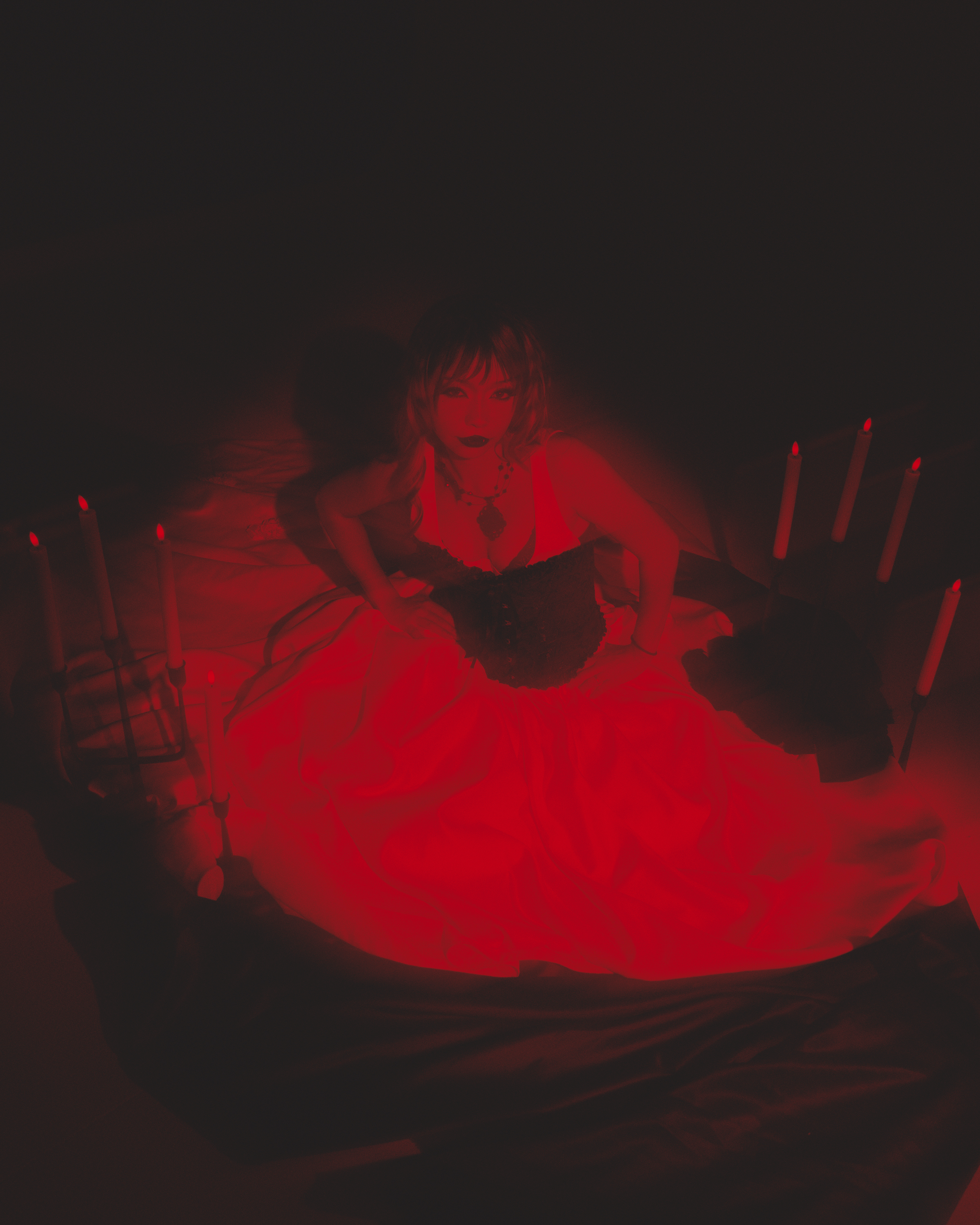 A woman dressed in gothic style with dark makeup and jewelry, sitting on the floor in a dark room illuminated by red lighting, with red candles around her.