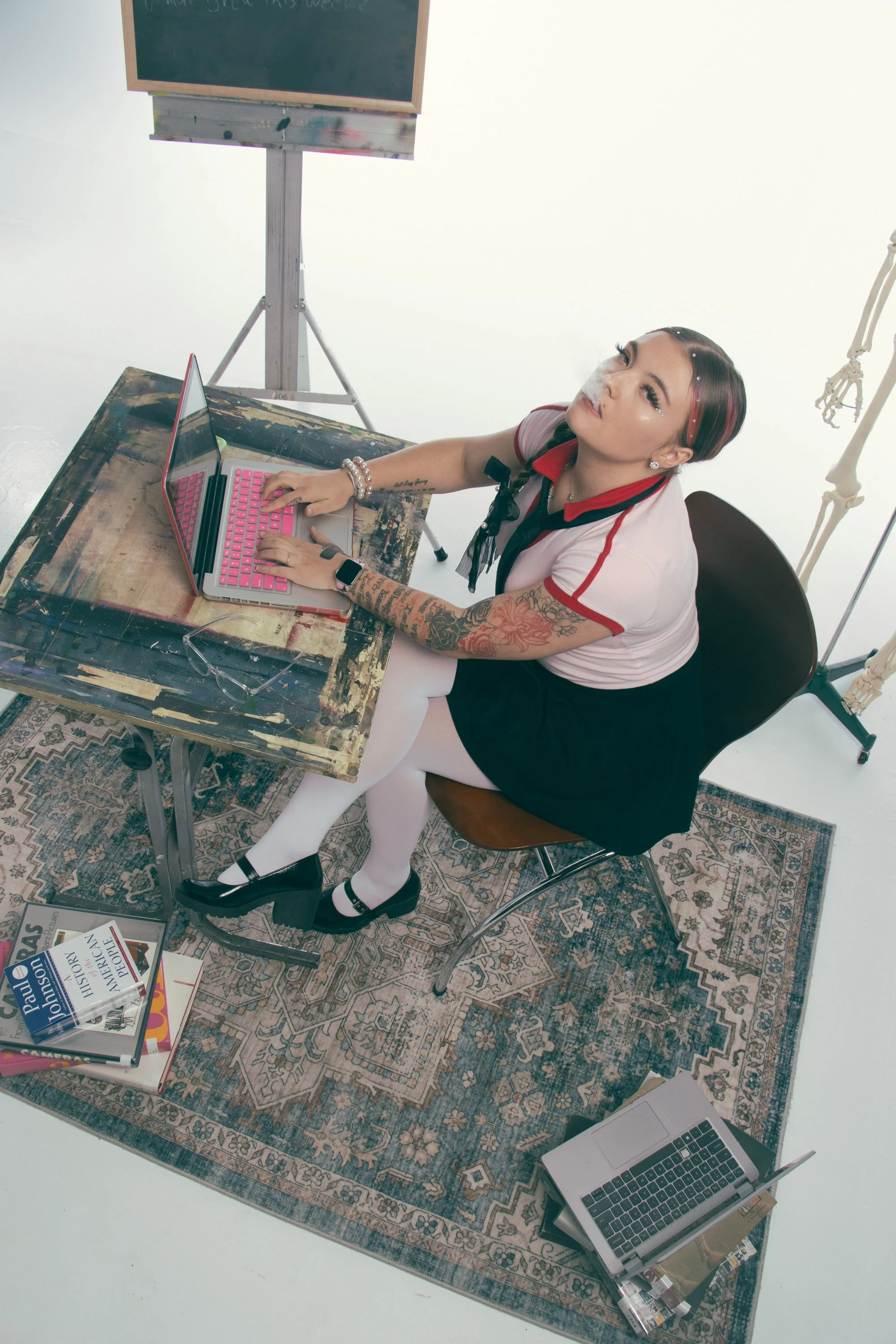 A young woman with tattoos, wearing white stockings, black shoes, a pearl bracelet, and a smartwatch, sitting at a wooden desk with a pink keyboard laptop. She has dark hair with red streaks and is exhaling smoke while looking up. The scene includes 