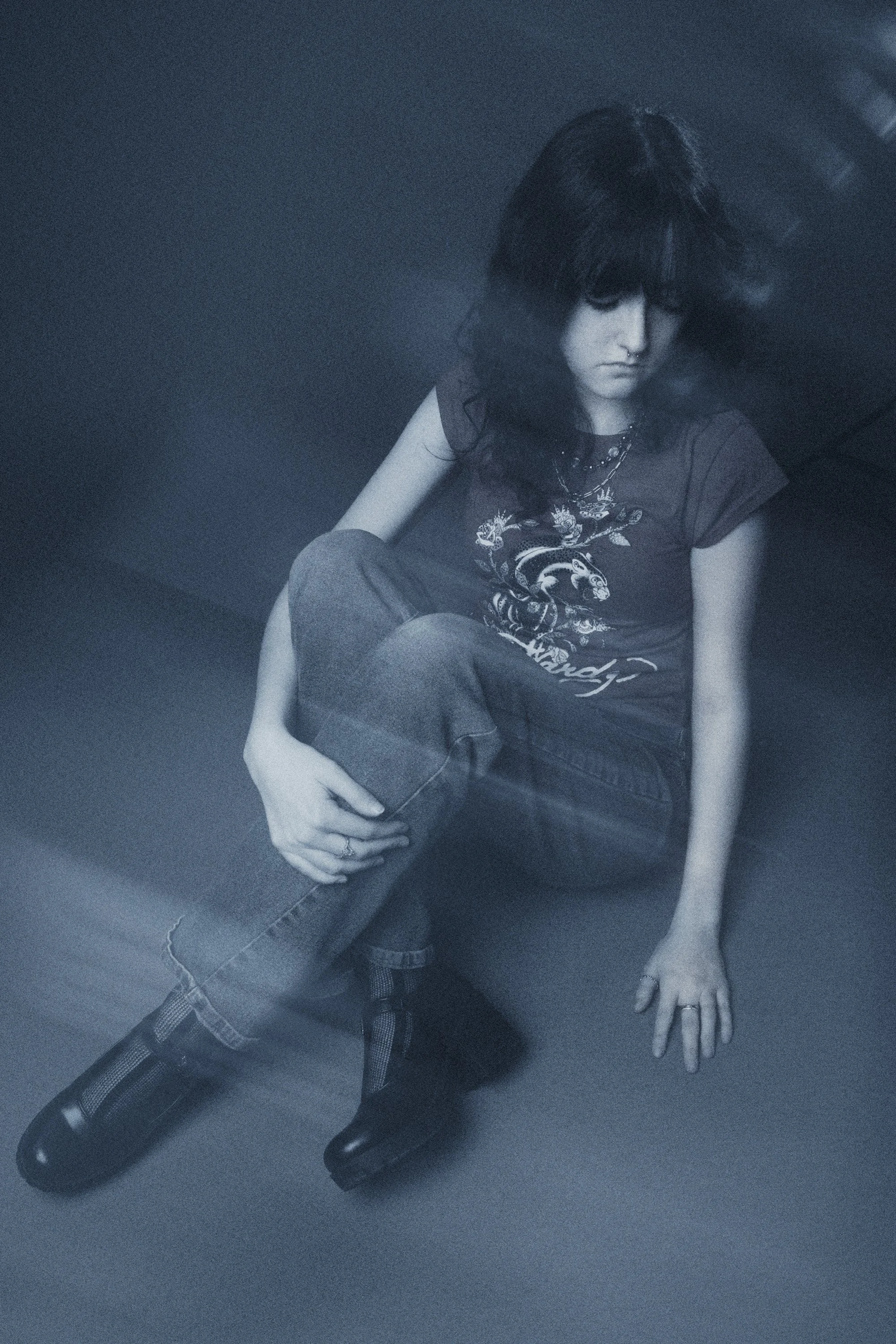 A woman with dark hair sitting on a dark floor, wearing jeans, a dark T-shirt with a dragon design, and boots. Her head is tilted downward and her left hand rests on her knee, with her right hand on the floor.